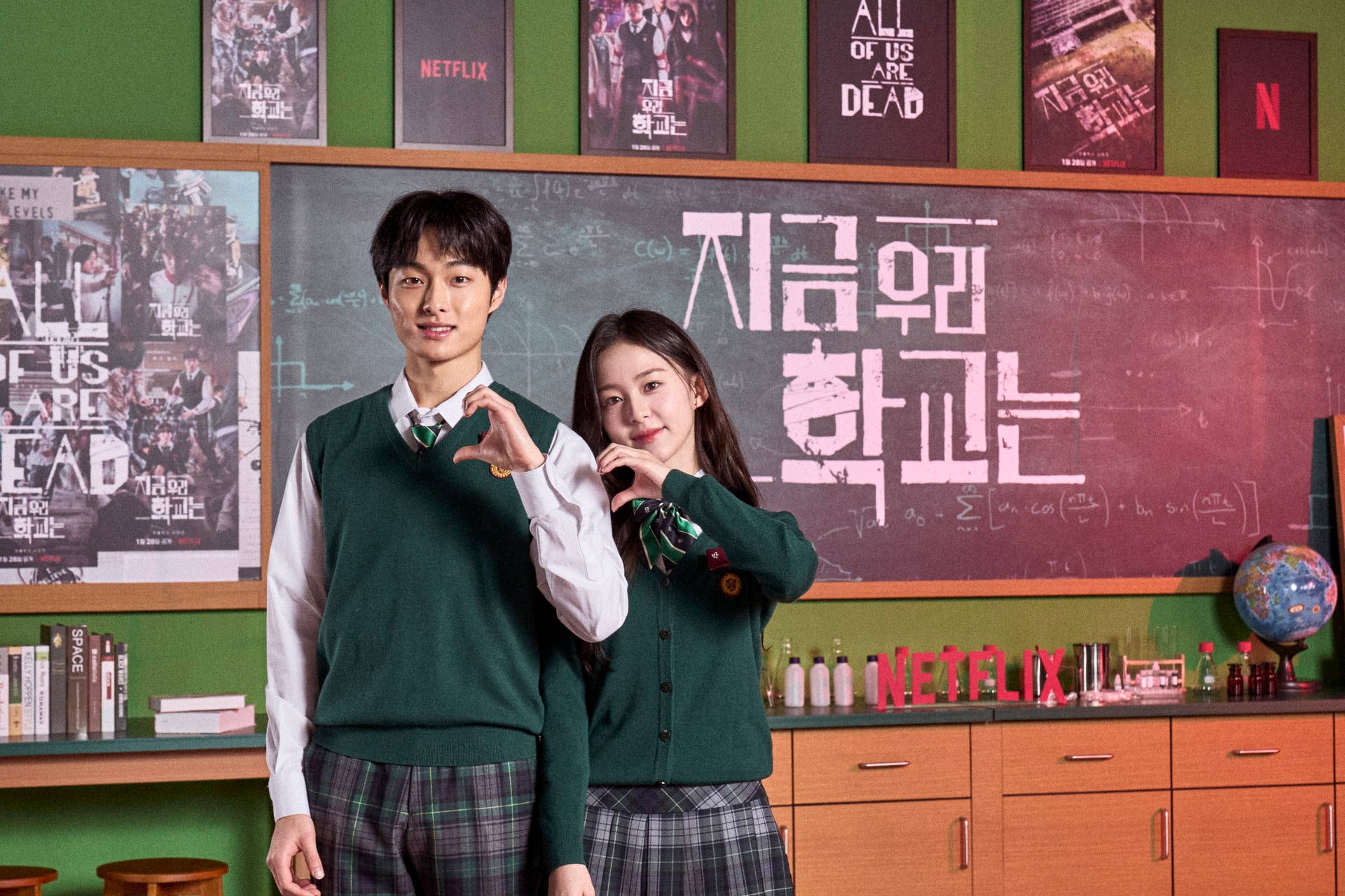 恐怖 桌布 壁紙 — 兩名穿著綠色校服的學生在佈滿韓文和Netflix殭屍劇海報的黑板前擺出心形手勢。