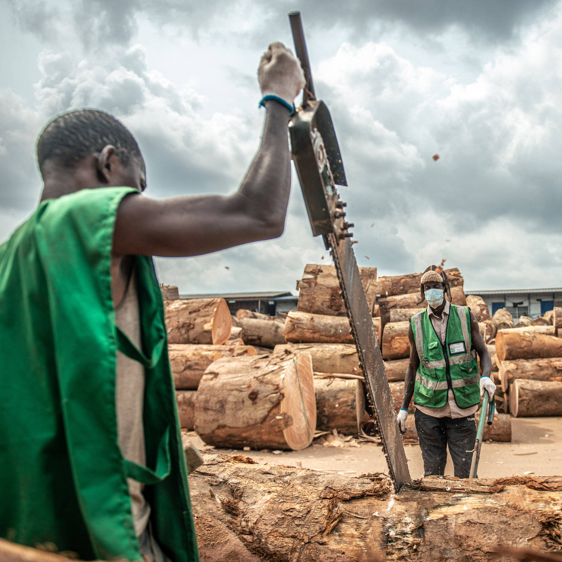 Africa 桌布 壁紙 — 兩名穿著綠色背心的工人在陰天下的木材場用鏈鋸切割原木。