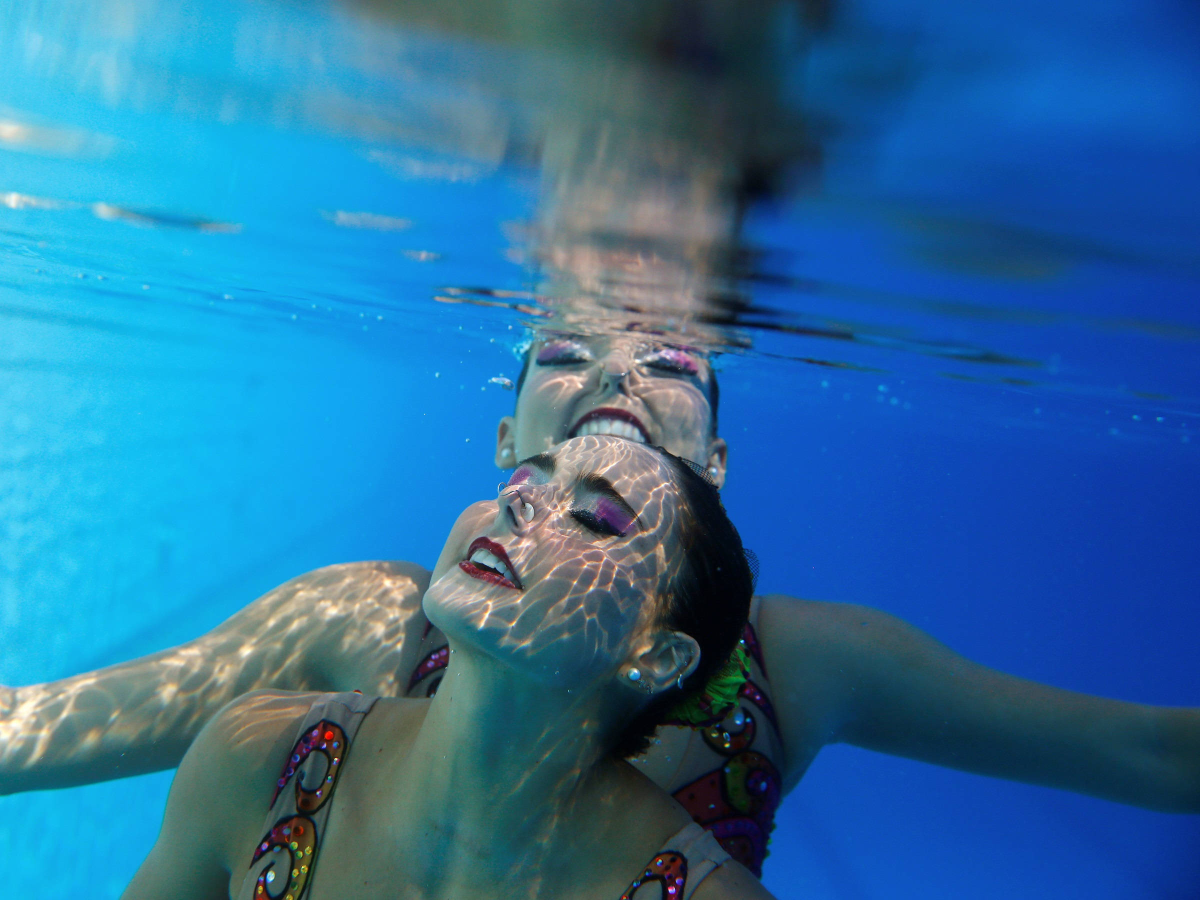 Swimming 桌布 壁紙 — 兩名穿著色彩繽紛泳衣的韻律泳選手在水中伸展雙臂滑行，陽光穿過清澈的藍色泳池水照射下來。