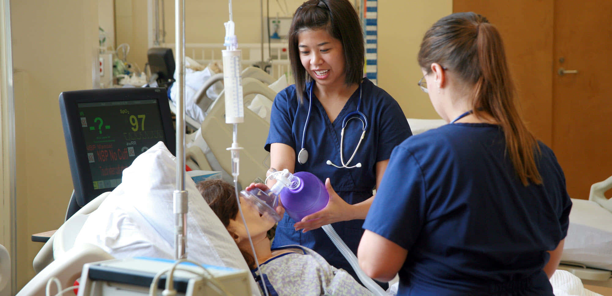 Nursing 桌布 壁紙 — 兩名穿著藍色工作服的微笑護士正在醫院病床旁為病人提供氧氣，附近有醫療監測儀器。