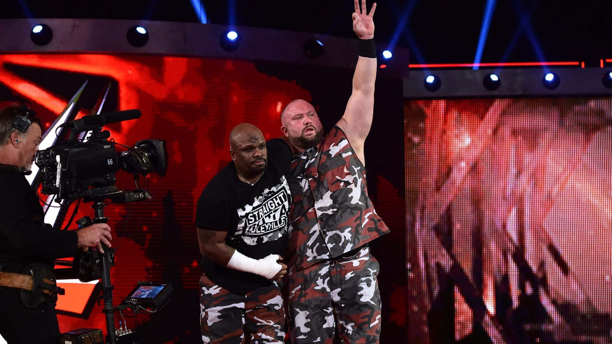 摔角 桌布 壁紙 — 兩名穿著迷彩裝備的摔角手在燈光璀璨的舞台上擺姿勢，一名攝影師正在拍攝他們。
