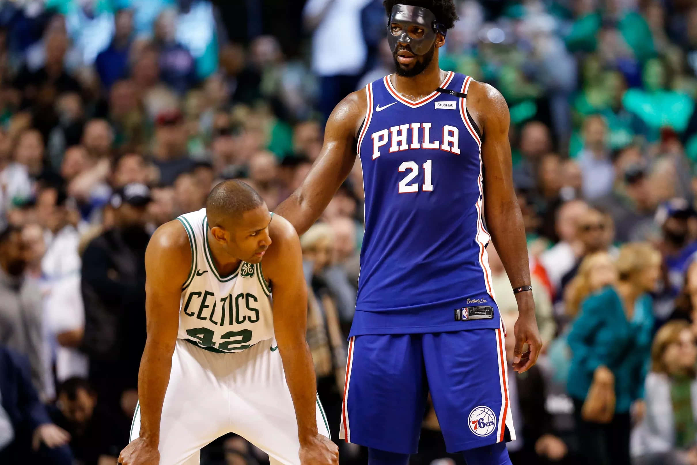 Al Horford 桌布 壁紙 — 兩名籃球運動員在比賽中對峙，一人身穿藍色費城球衣並戴著口罩，另一人身穿白色波士頓塞爾提克球衣。
