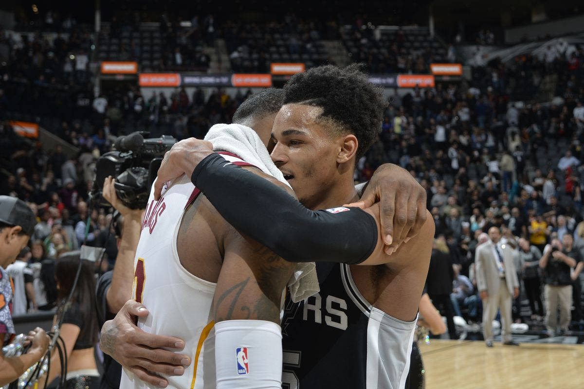 Dejounte Murray 桌布 壁紙 — 兩名籃球運動員在燈光明亮的球場上熱情擁抱，背景是座無虛席的體育館。