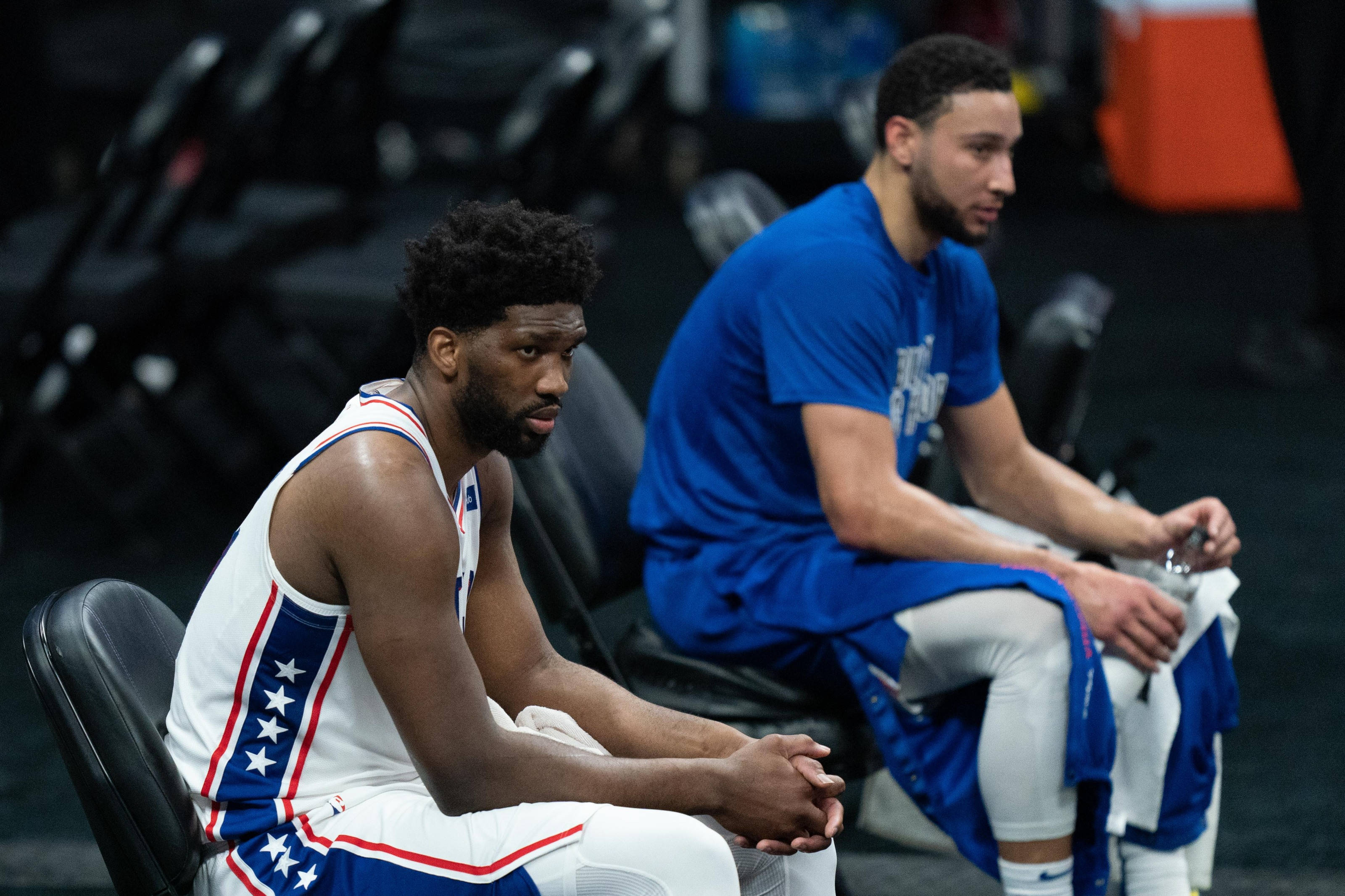 76ers 桌布 壁紙 — 兩名籃球運動員坐在球場的黑色椅子上，一人身穿白色星星球衣，另一人身穿藍色熱身服，兩人都專注而嚴肅。