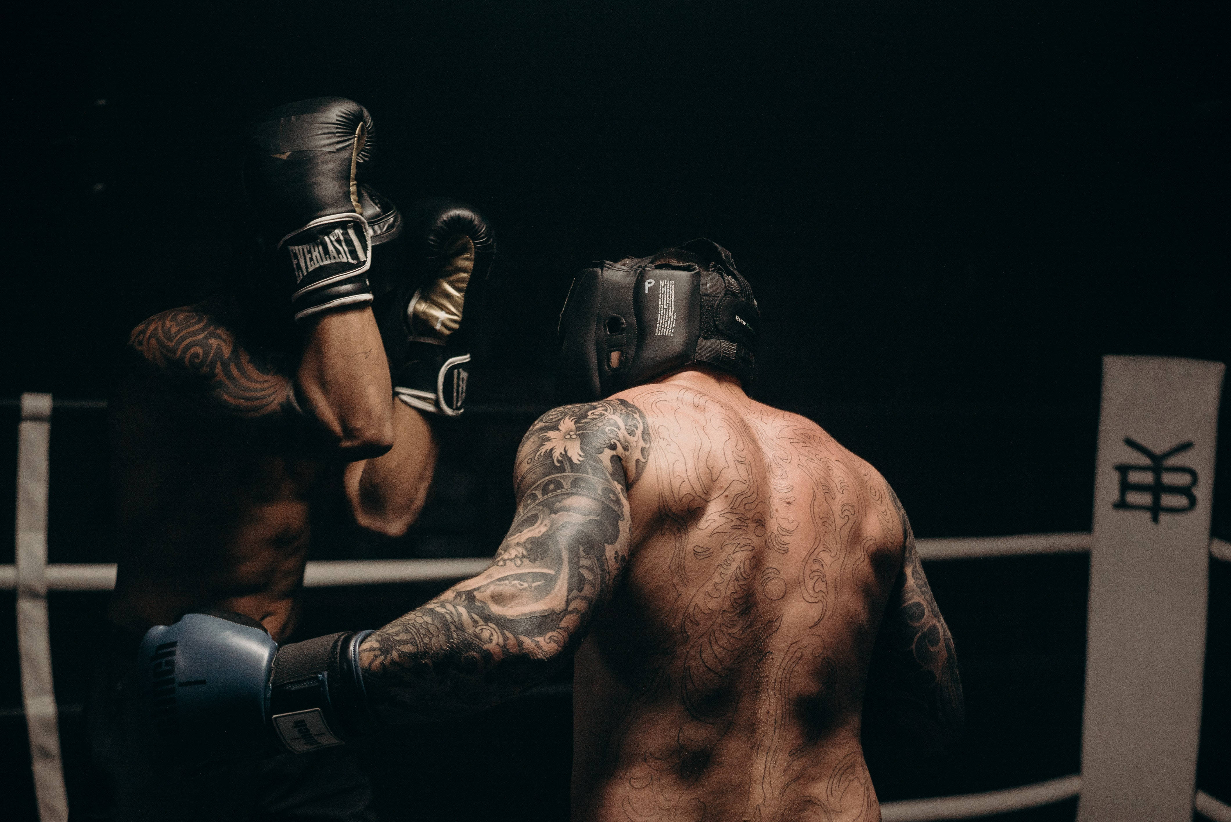 Cool Boxing 桌布 壁紙 — 兩名紋身拳擊手在燈光昏暗的擂台上交鋒，其中一人揮動戴手套的拳頭擊向對方的頭部。