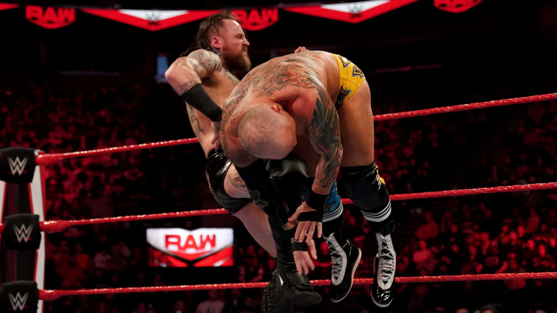 WWE 桌布 壁紙 — 兩名紋身摔角手在 RAW 品牌下，於燈光明亮、有紅色繩索和喧鬧人群的擂台上激烈纏鬥。