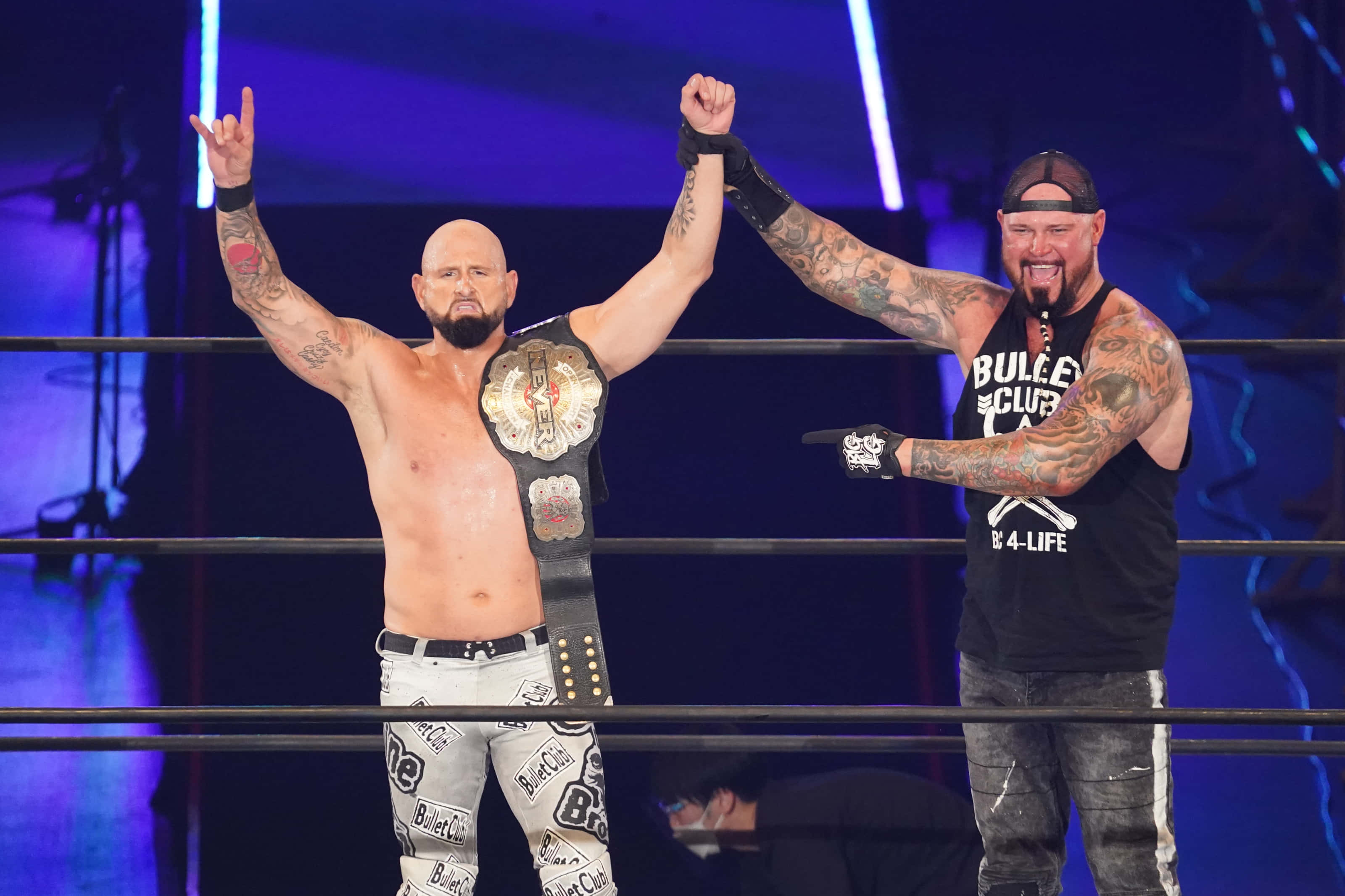 WWE 桌布 壁紙 — 兩名紋身摔角手在擂台上慶祝，一人手持冠軍腰帶，另一人在藍色舞台燈光下舉起拳頭。