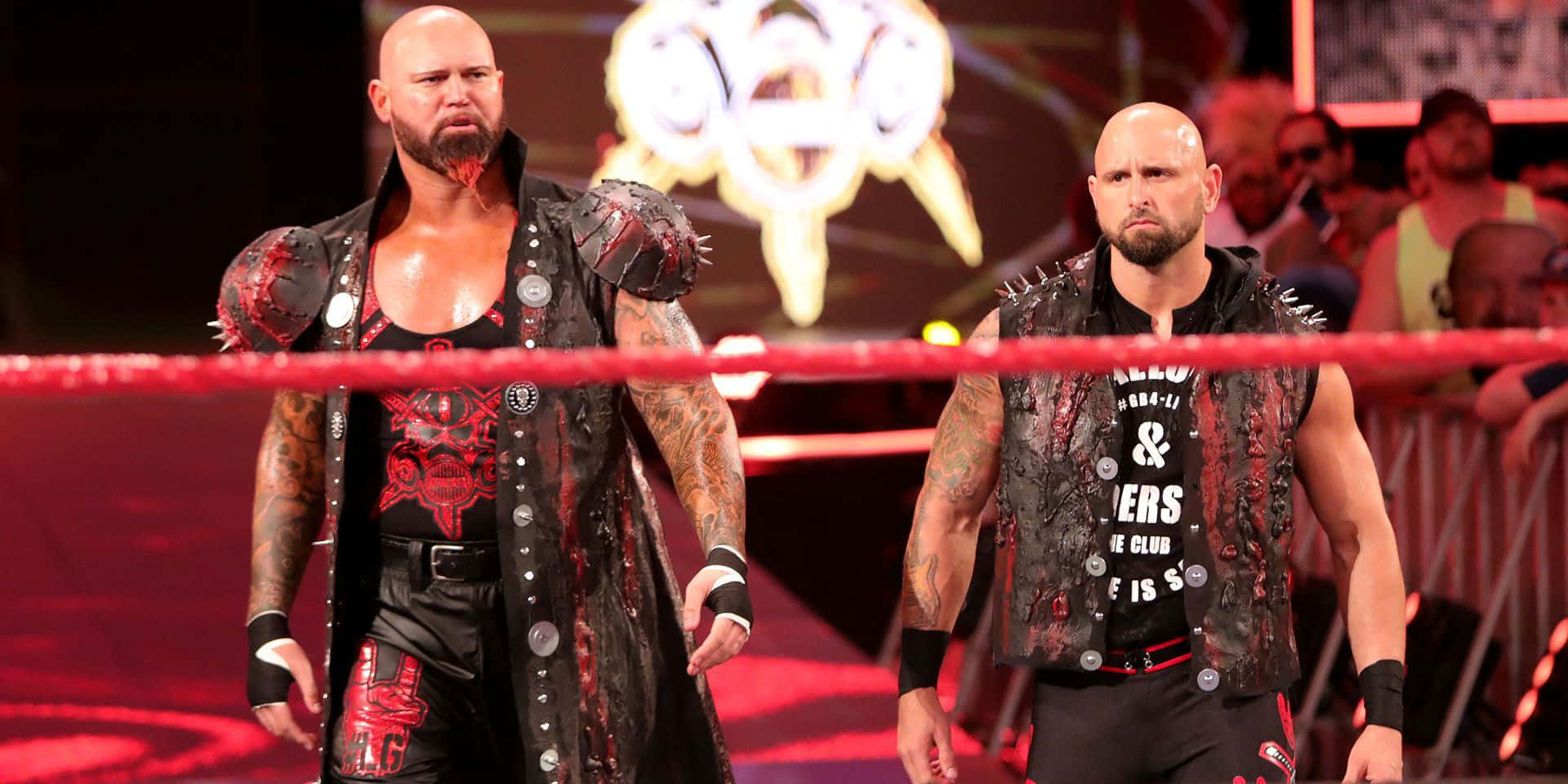 WWE 桌布 壁紙 — 兩名紋身濃密的光頭摔角手，身穿黑色尖刺裝備，在明亮的場館燈光下，威嚇地站在紅色擂台上。