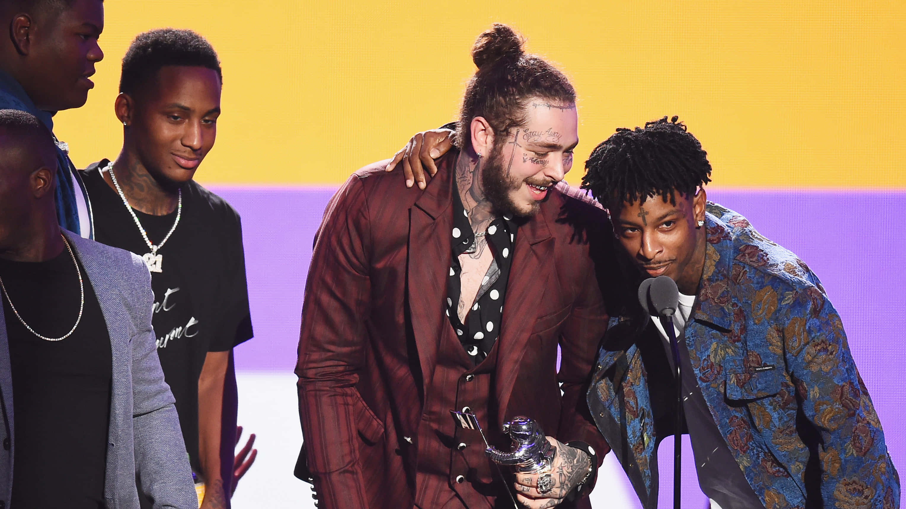 21 Savage 桌布 壁紙 — 兩名紋身男子穿著時尚西裝，在燈光明亮的舞台上分享歡樂時刻，背景為黃色和紫色。