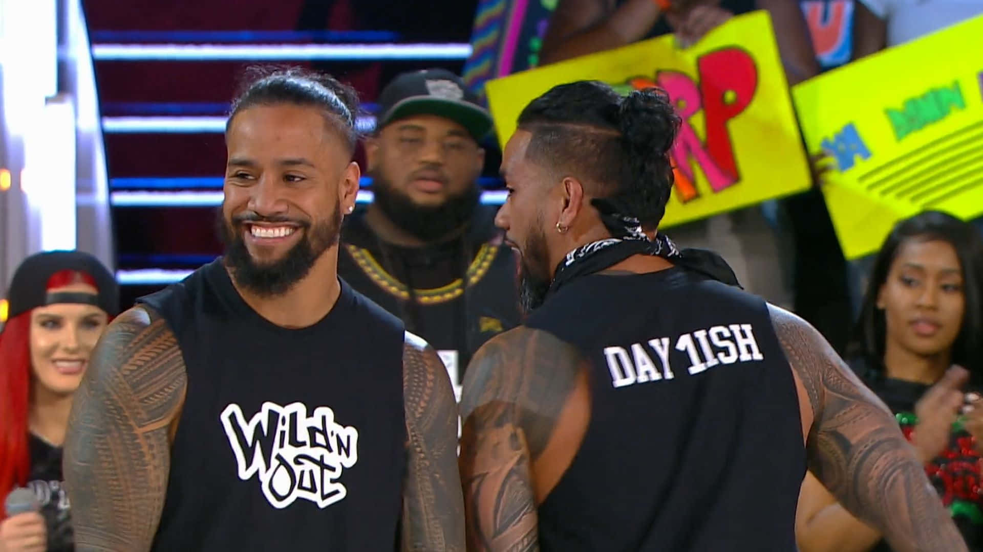 Jey Uso 桌布 壁紙 — 兩名紋身男子身穿黑色背心，在燈光璀璨的舞台上微笑互動，身後有粉絲舉著標語牌。