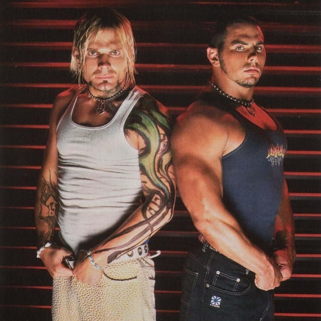 WWE 桌布 壁紙 — 兩名紋身肌肉男在粗獷的攝影棚環境中，自信地以紅色條紋背景擺姿勢。