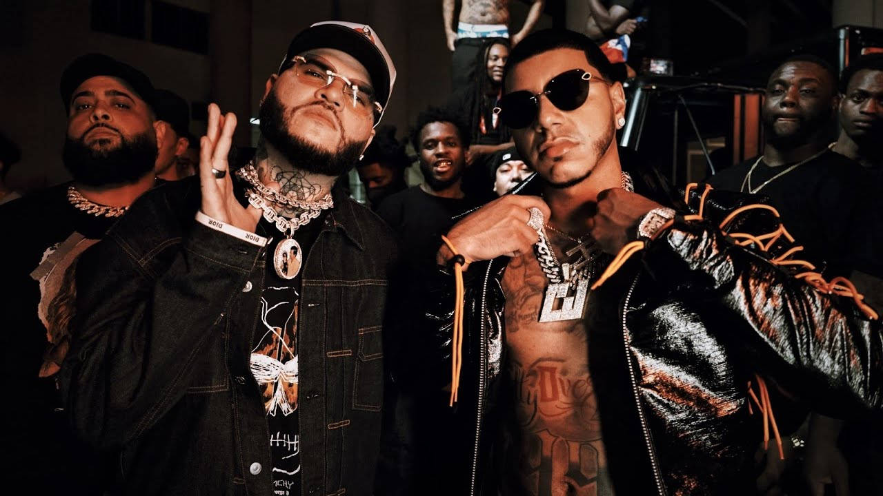 farruko 桌布 壁紙 — 兩名紋身說唱歌手身穿黑色皮夾克和鏈條，在燈光昏暗的音樂會上自信地擺姿勢，身後有粉絲。