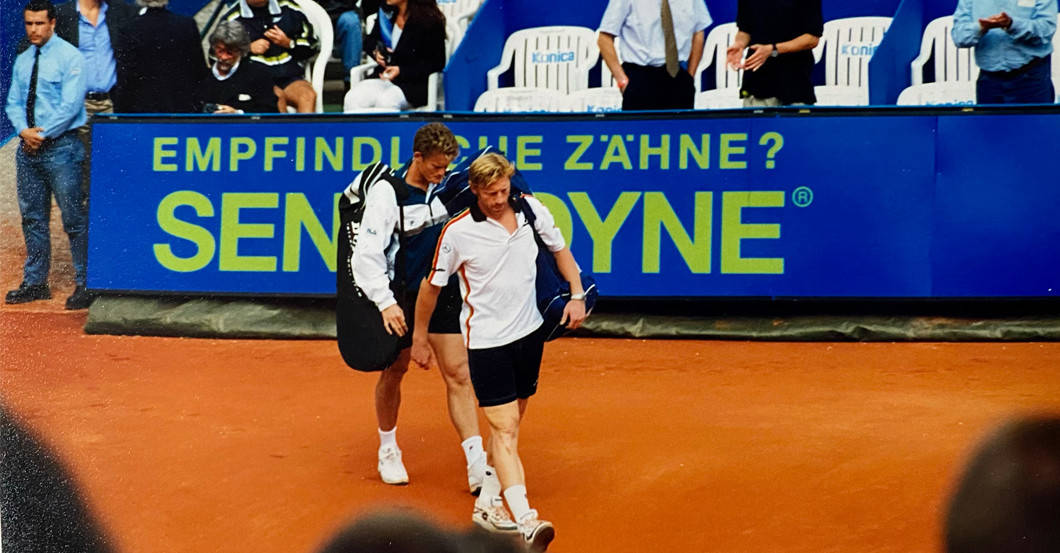 Tennis 桌布 壁紙 — 兩名網球運動員在比賽後離開紅土球場，一人攙扶另一人，背景有觀眾和藍色贊助商橫幅。