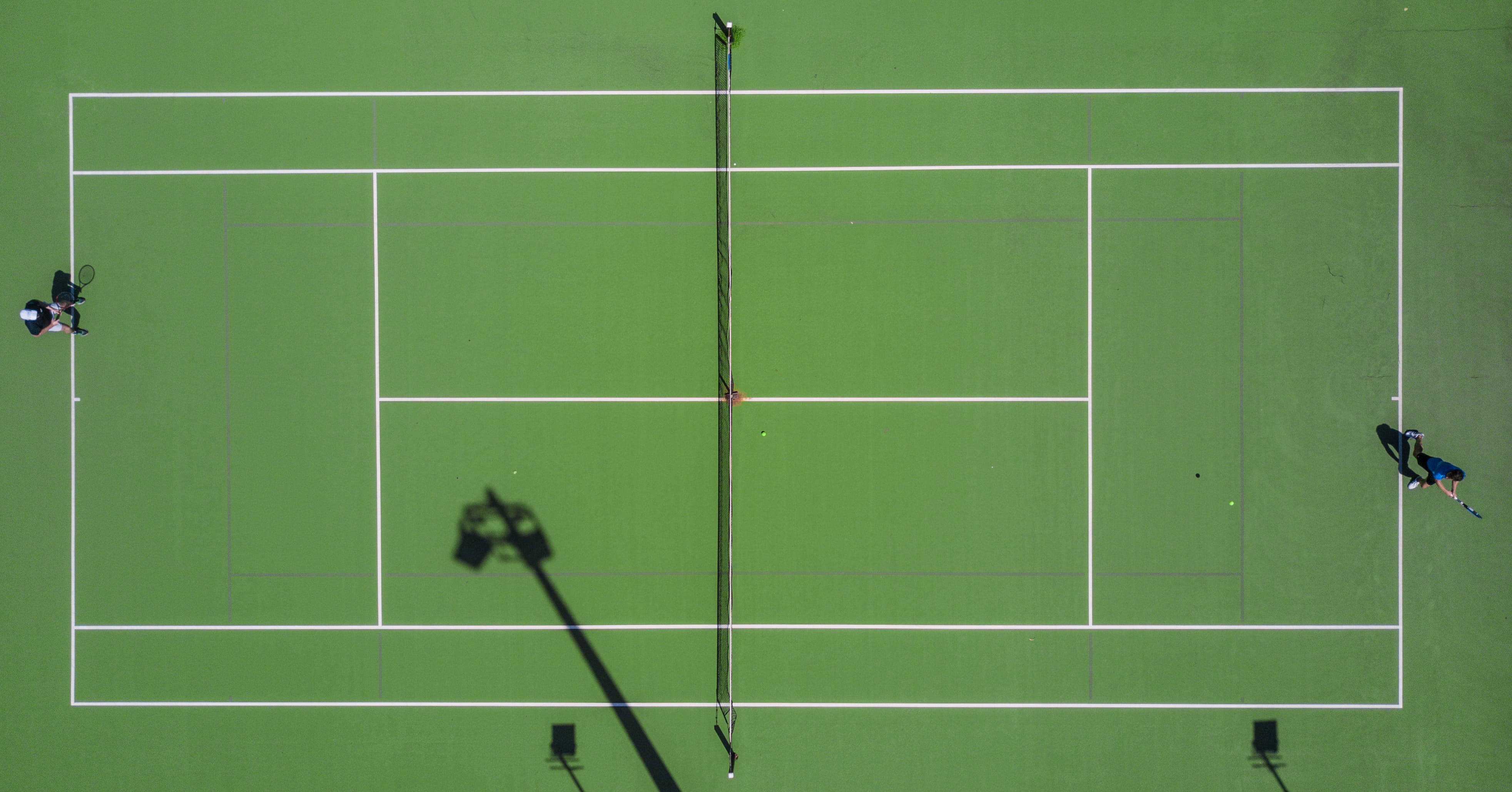 Wimbledon 桌布 壁紙 — 兩名網球運動員在鮮綠色球場上於明亮日光下打球，帶有長影的鳥瞰圖。