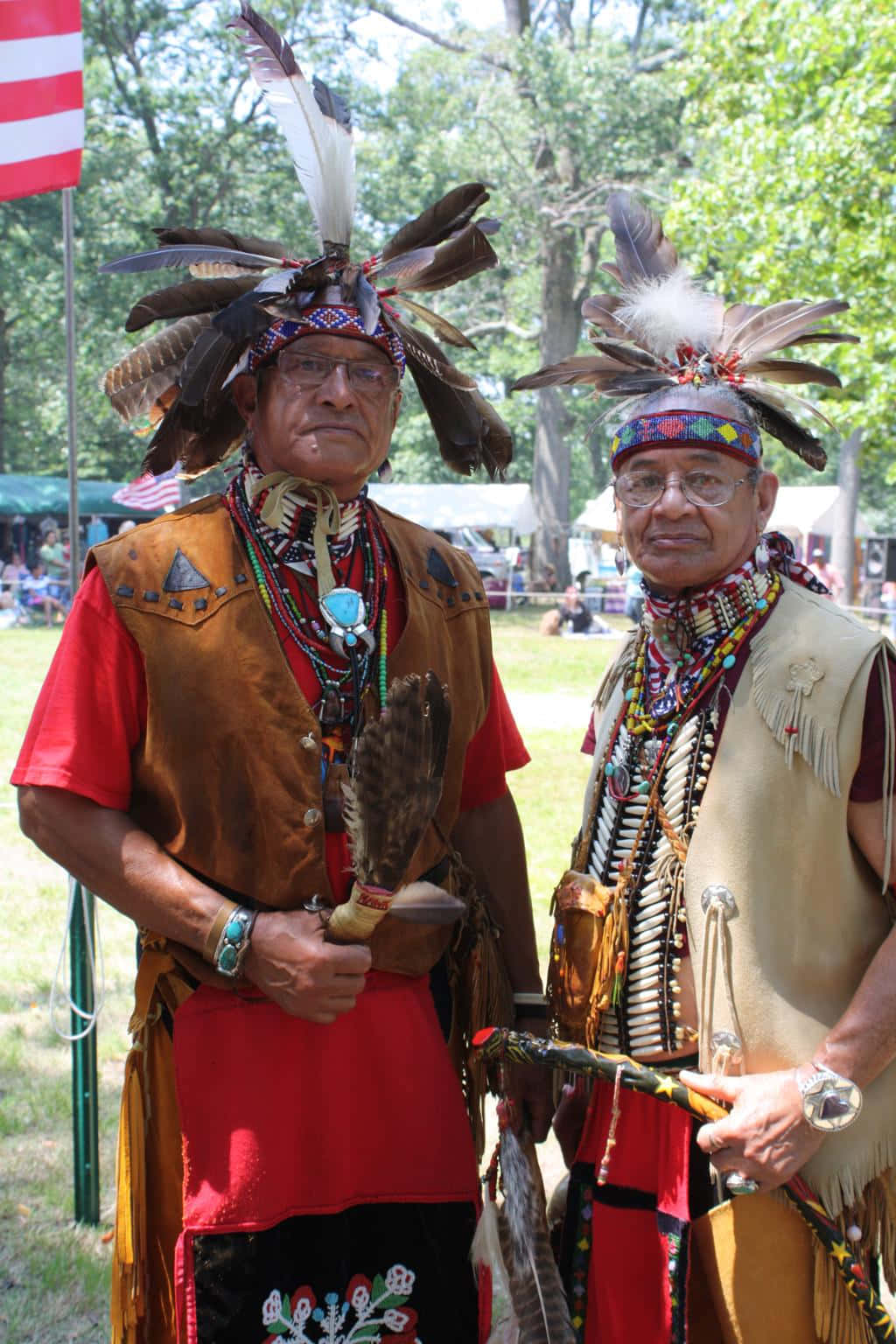 Native American 桌布 壁紙 — 兩名美洲原住民男子身穿傳統羽毛頭飾和珠飾背心，在明亮的日光下莊嚴地站在戶外文化聚會中。