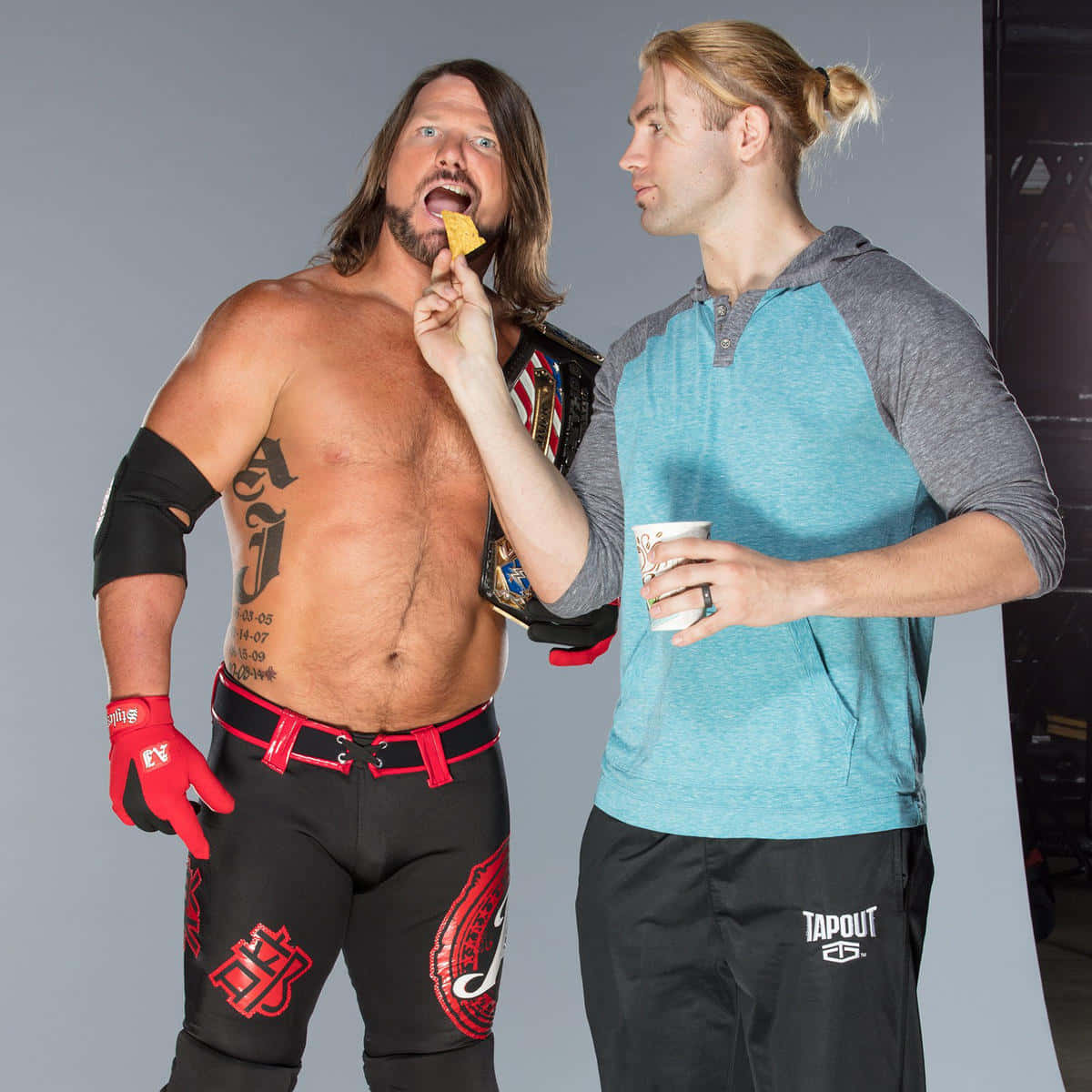 WWE 桌布 壁紙 — 兩名肌肉男，一人身穿摔角裝備正在吃薯片，另一人身穿休閒服裝拿著杯子觀看。