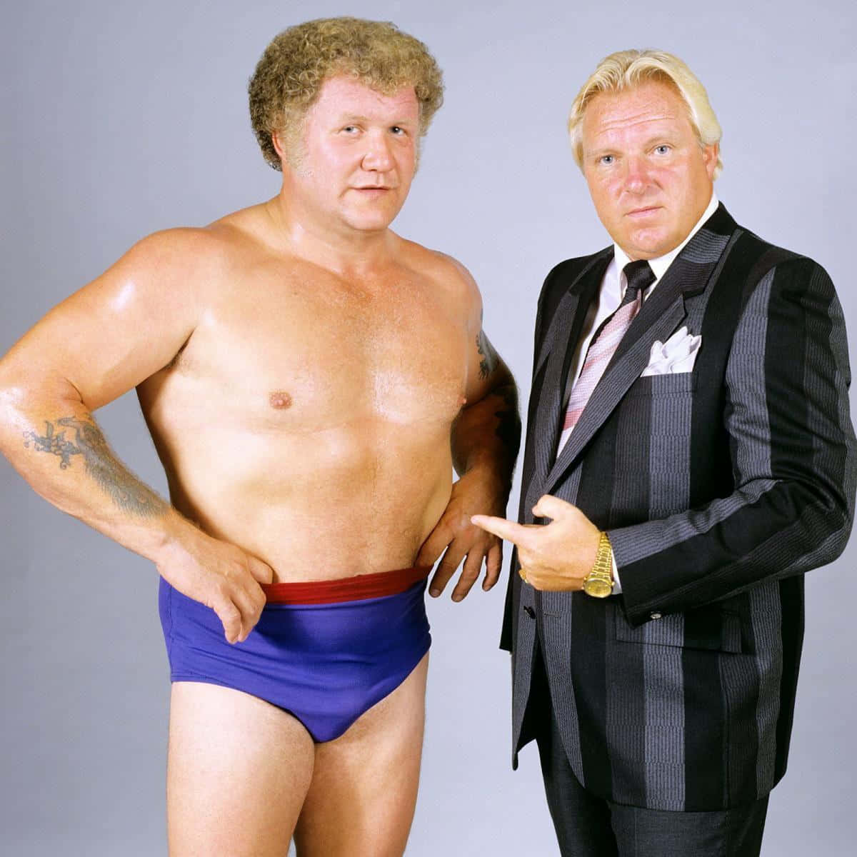 WWE 桌布 壁紙 — 兩名肌肉男在灰色背景前擺姿勢，一人穿藍色摔角褲，另一人穿細條紋西裝指著他。
