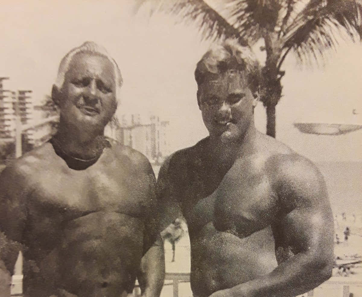WWE 桌布 壁紙 — 兩名肌肉男在陽光明媚的海灘上赤裸上身擺姿勢，背景有棕櫚樹和遠處的建築物，是一張復古顆粒感的照片。