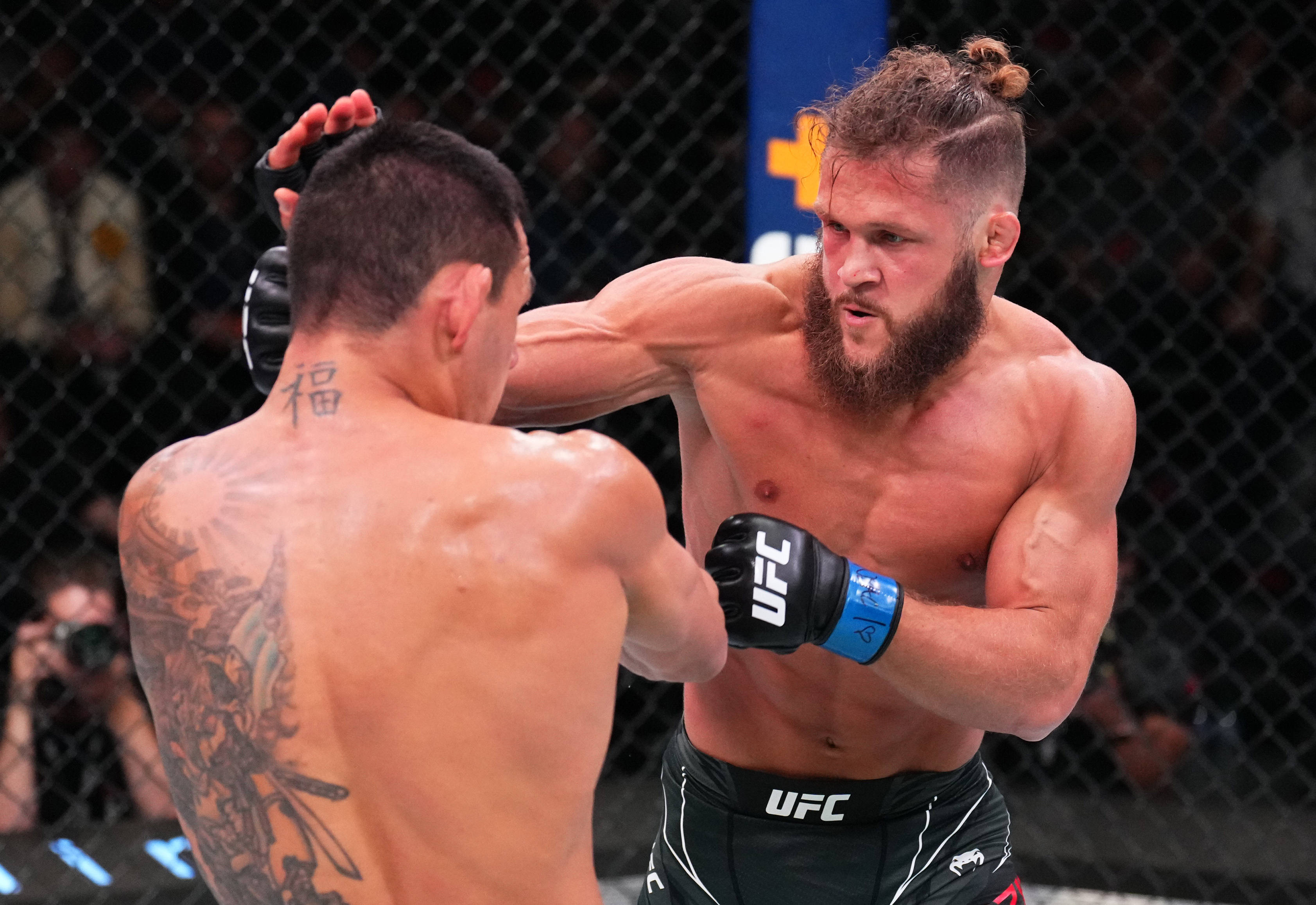 UFC 桌布 壁紙 — 兩名肌肉發達的MMA拳手在激烈的鐵籠賽中交鋒，汗水飛濺，紋身在明亮的場館燈光下清晰可見。