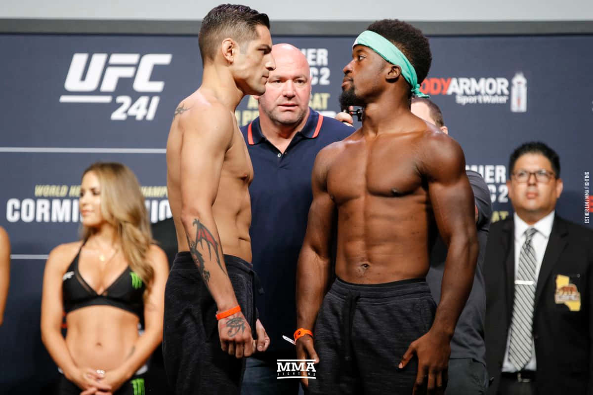MMA 桌布 壁紙 — 兩名肌肉發達的UFC拳手在稱重活動中激烈對峙，背景有官員和一名比基尼模特兒。