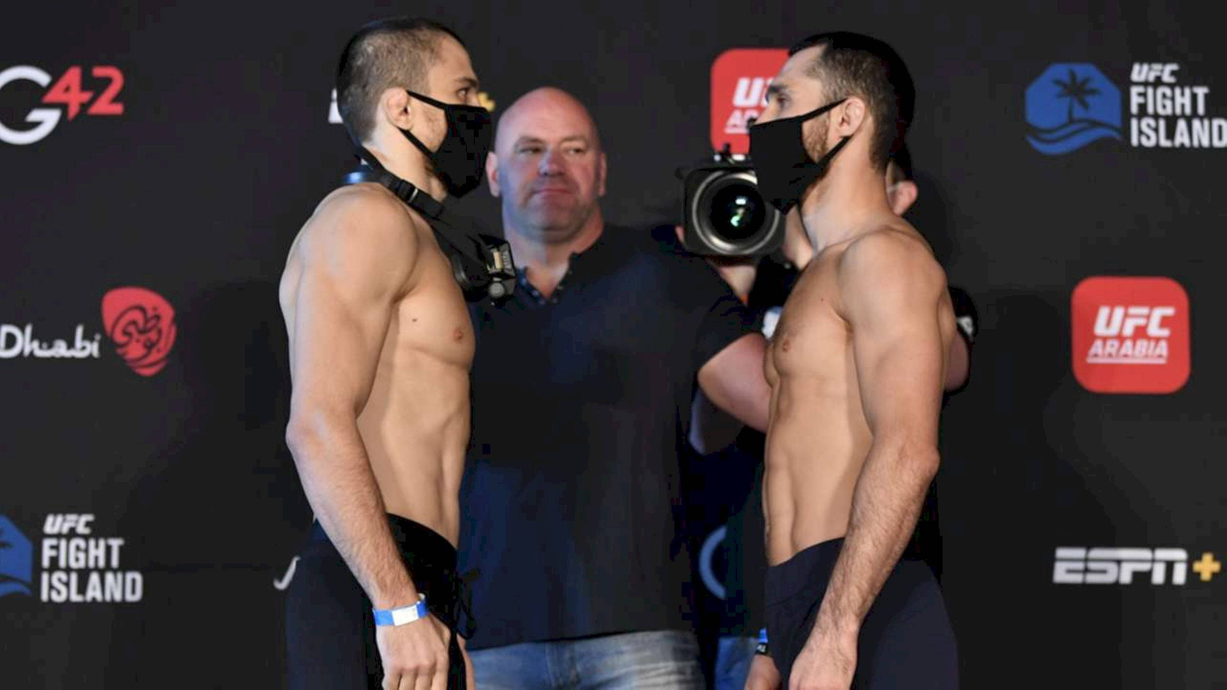 UFC 桌布 壁紙 — 兩名肌肉發達的UFC拳手在稱重活動中戴著口罩對峙，背景有裁判和攝影團隊。