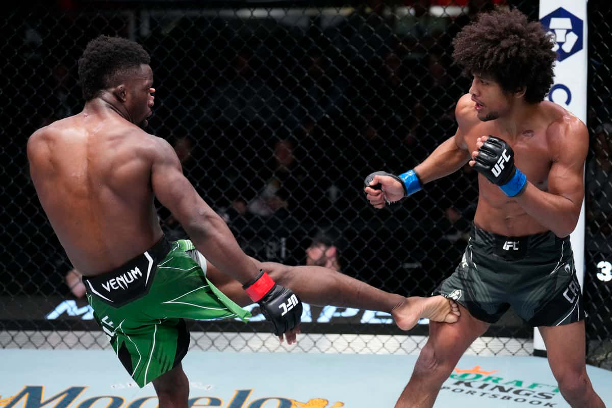 UFC 桌布 壁紙 — 兩名肌肉發達的拳手在UFC八角籠中交鋒，一人使出高踢，另一人在明亮的場館燈光下格擋。