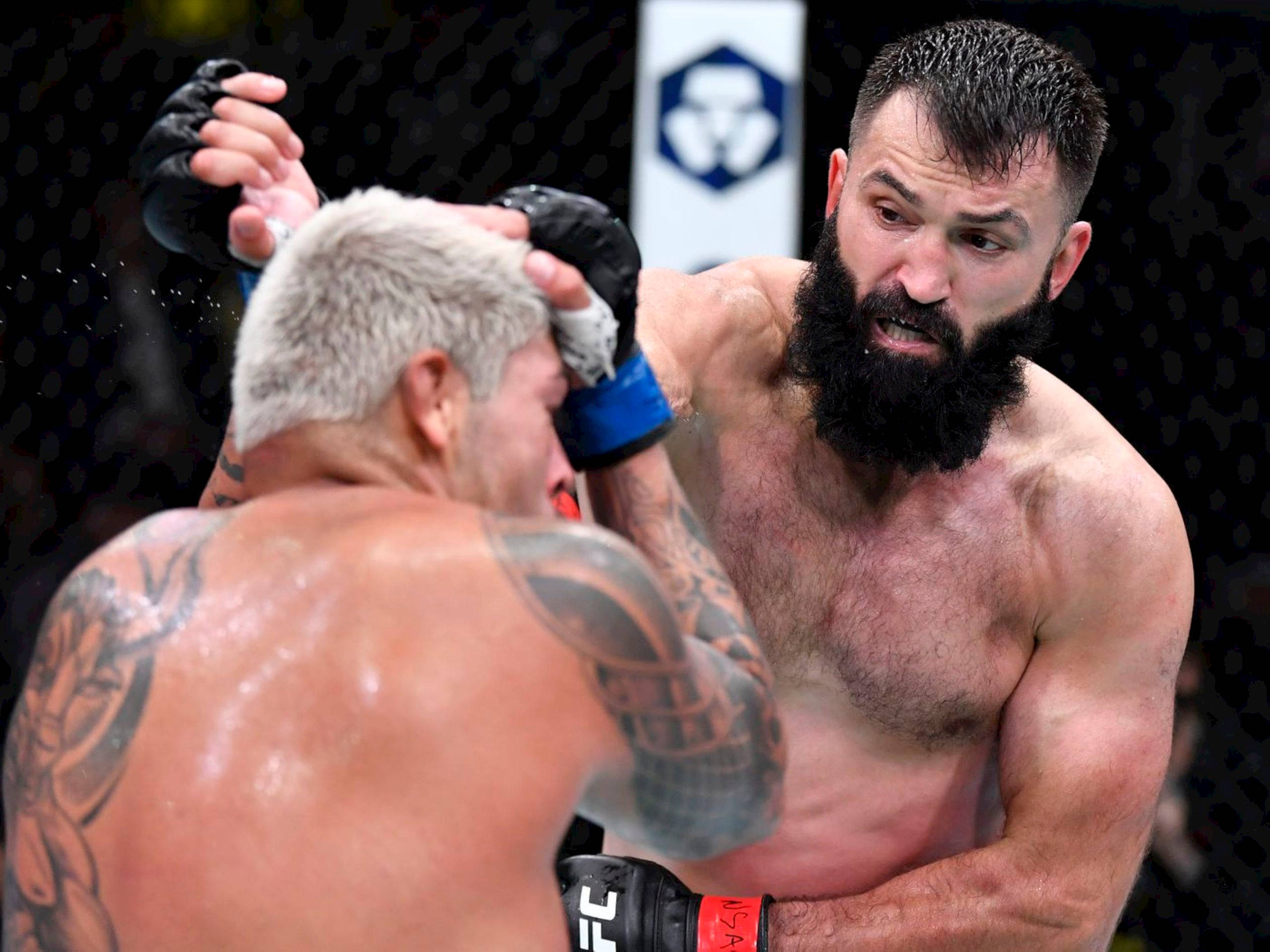 UFC 桌布 壁紙 — 兩名肌肉發達的拳手在UFC八角籠中交鋒，其中一人擊中對方頭部，汗水在激烈戰鬥中飛濺。