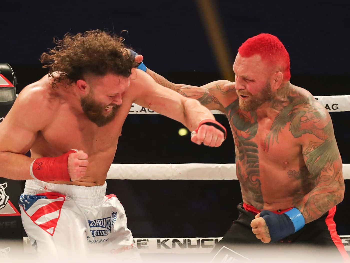 MMA 桌布 壁紙 — 兩名肌肉發達的拳手在拳擊場上交鋒，一名紅髮紋身的拳手向他的對手（捲髮、穿美國國旗短褲）揮出一拳。