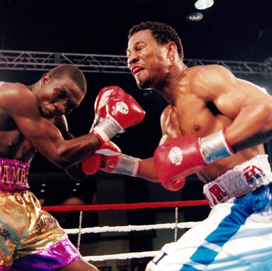 Boxing 桌布 壁紙 — 兩名肌肉發達的拳擊手在擂台上交鋒，其中一人在明亮的場館燈光下，用紅色拳套重擊對手的臉部。