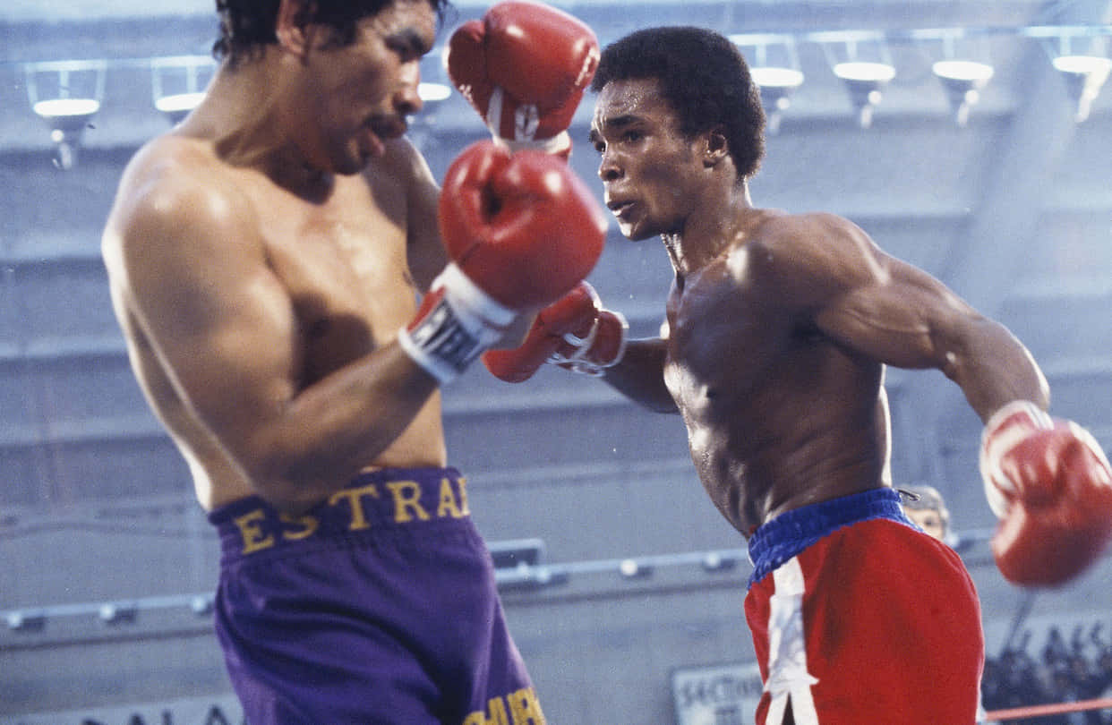Boxing 桌布 壁紙 — 兩名肌肉發達的拳擊手在緊張的擂台上碰撞，紅色手套和汗水在明亮的場館燈光下閃閃發光。