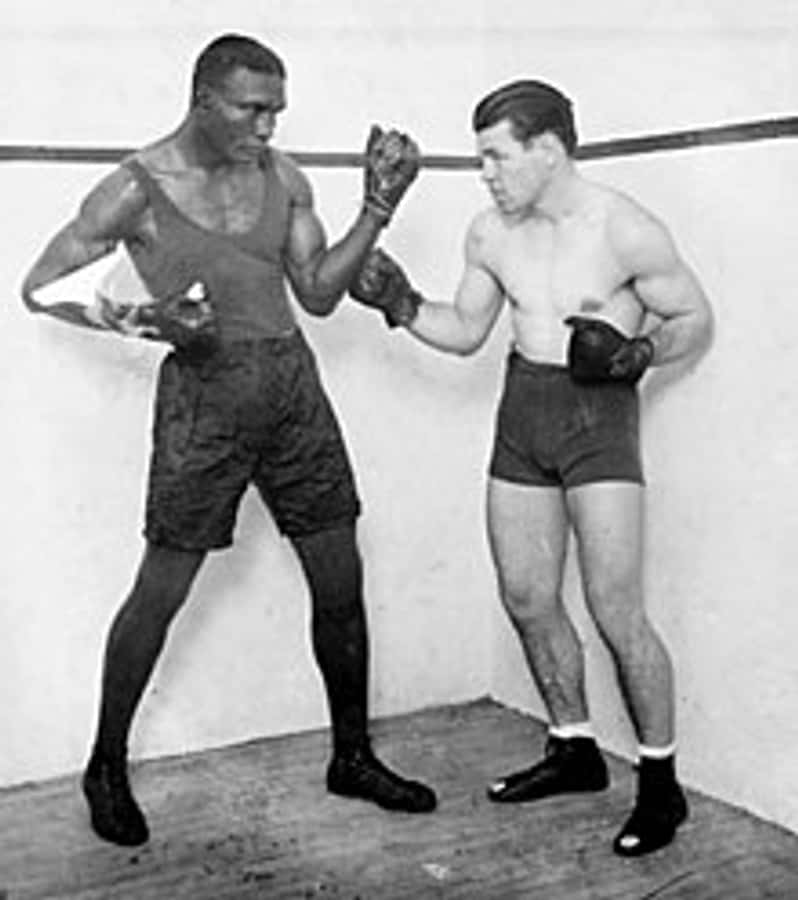 boxer 桌布 壁紙 — 兩名肌肉發達的拳擊手穿著復古裝備，在燈光昏暗的擂台上以專注的強度對峙。