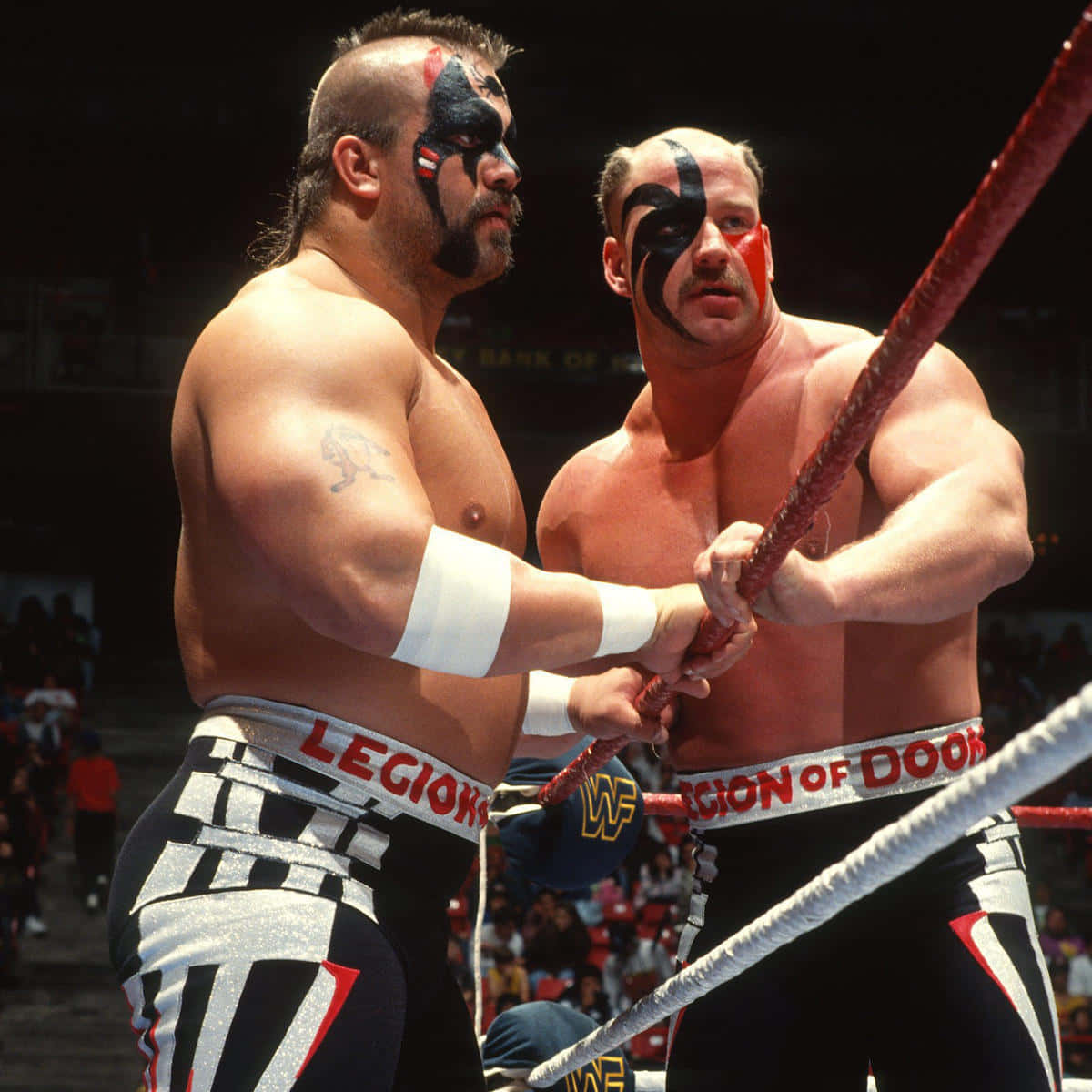 WWE 桌布 壁紙 — 兩名肌肉發達的摔角手，臉上塗著黑色和紅色的顏料，在明亮的競技場燈光下緊張對峙，抓住擂台繩索。