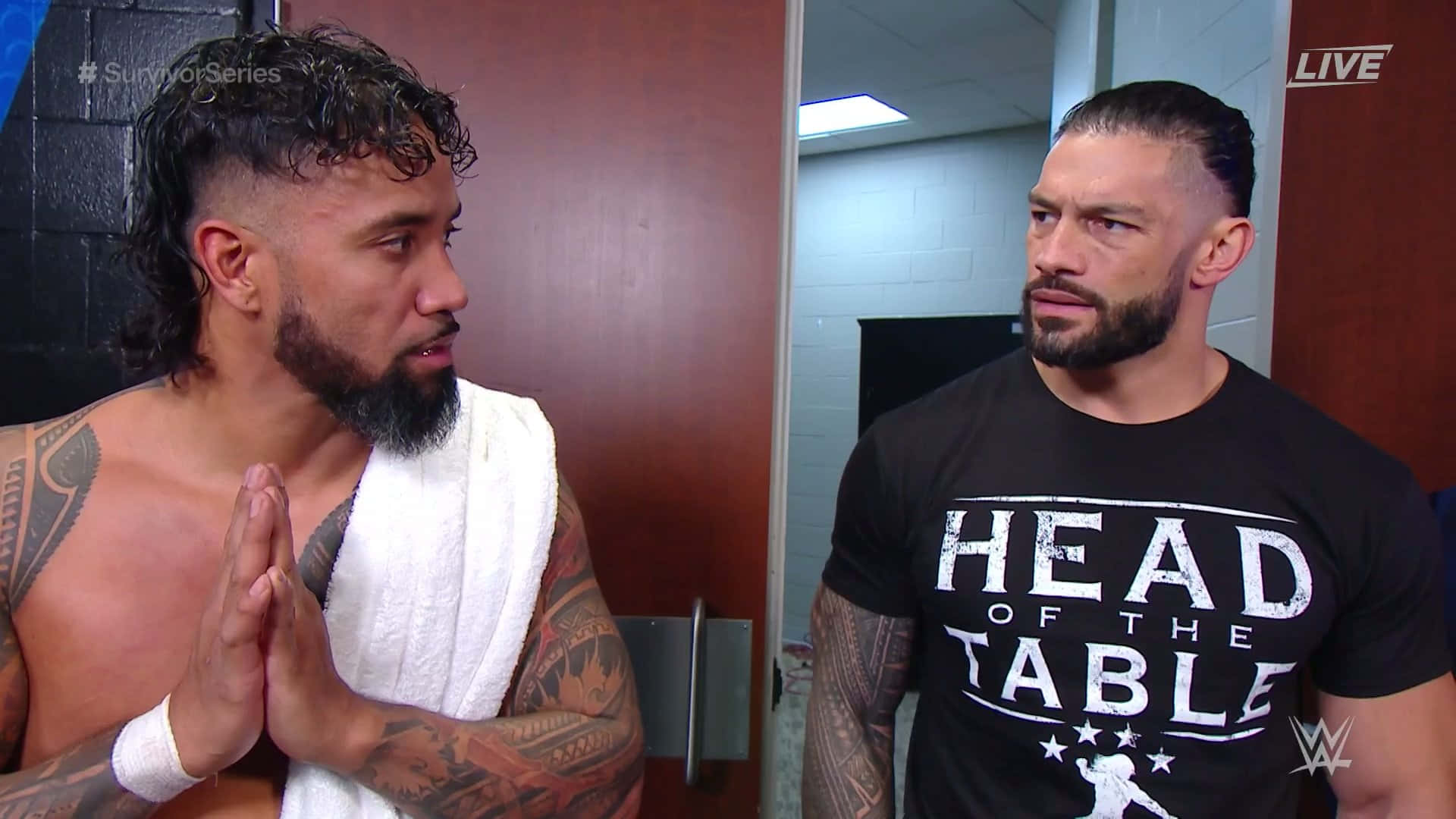 Roman Reigns 桌布 壁紙 — 兩名肌肉發達的摔角手在後台走廊對峙，一人赤裸上身披著毛巾，另一人穿著黑色「Head of the Table」T恤。
