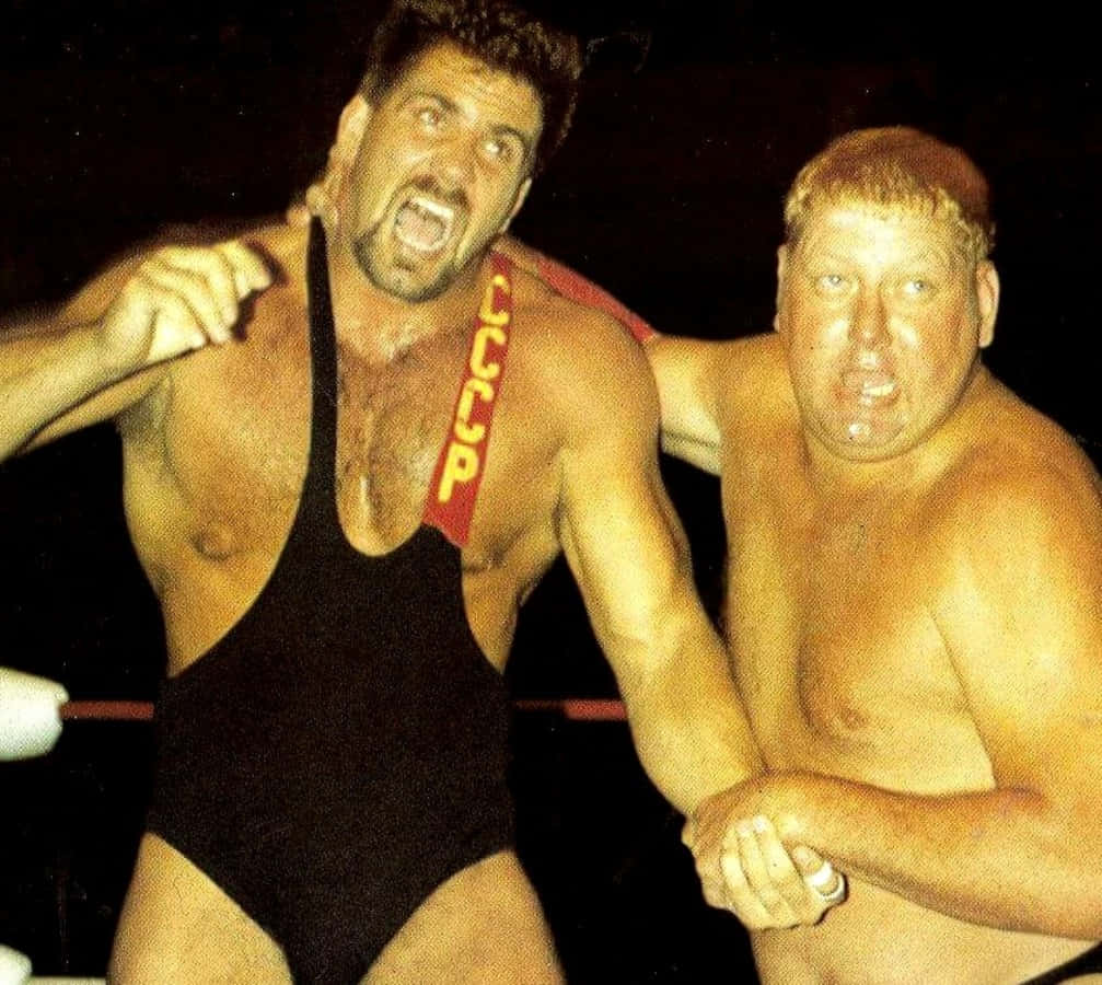 Dusty Rhodes 桌布 壁紙 — 兩名肌肉發達的摔角手在擂台上，一人身穿帶有紅色CCCP帶子的黑色緊身衣，另一人赤裸上身，兩人在明亮燈光下表情激烈。