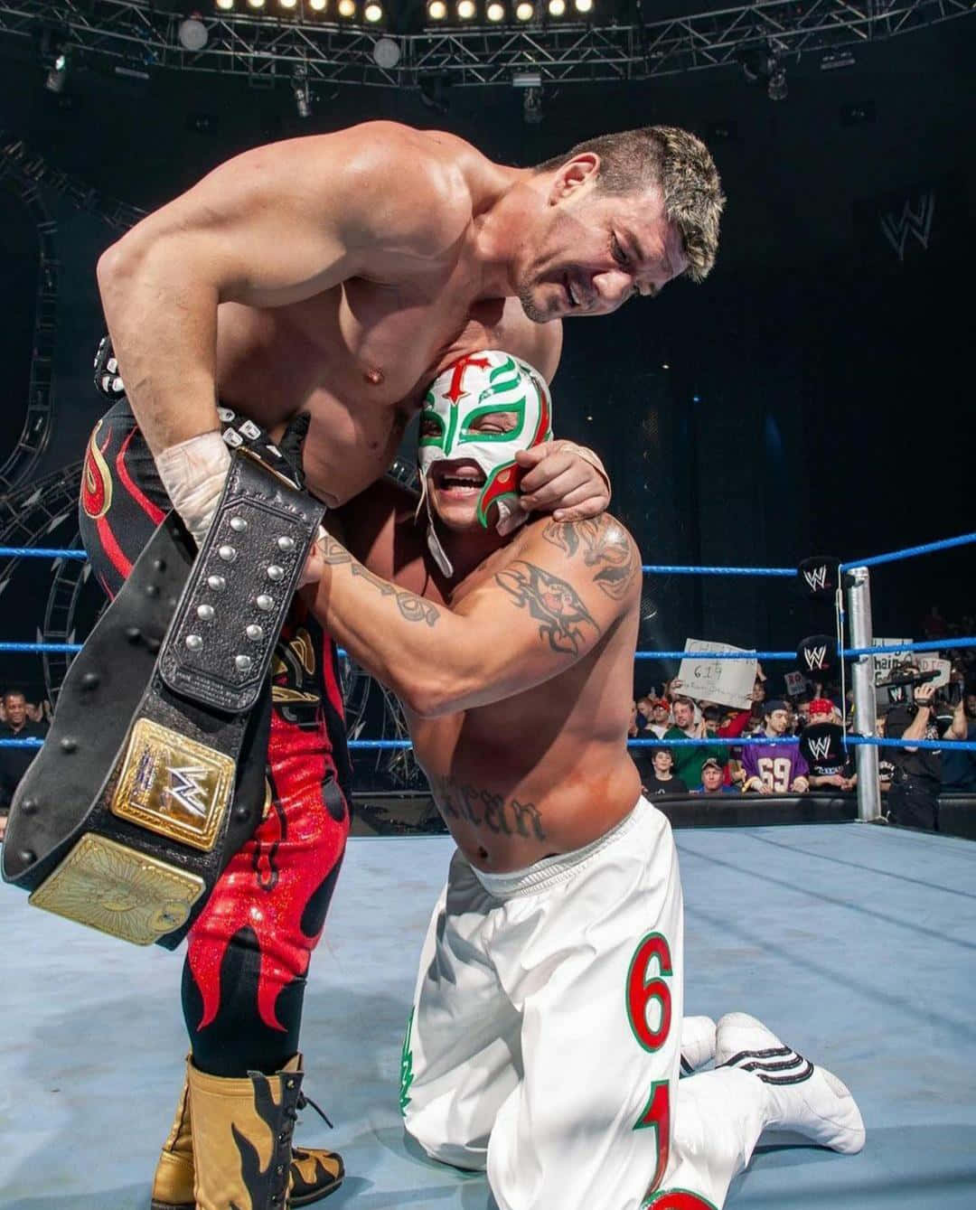 Wrestling 桌布 壁紙 — 兩名肌肉發達的摔角手在擂台上，其中一人手持冠軍腰帶，壓制著戴面具跪地認輸的對手，在明亮的場館燈光下。