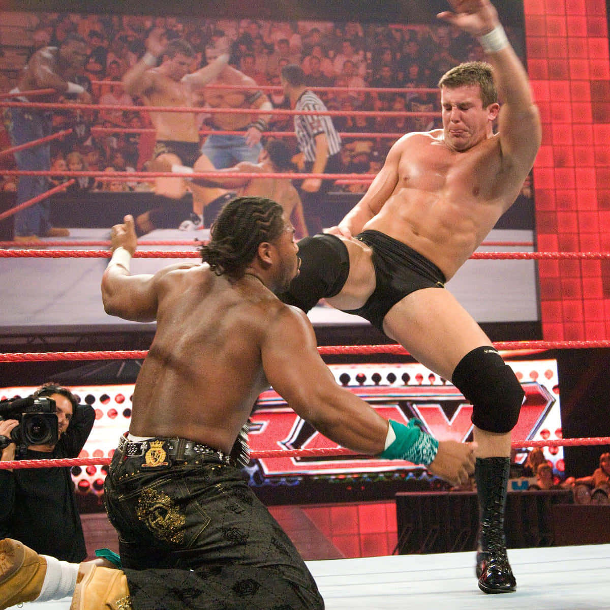 摔角 桌布 壁紙 — 兩名肌肉發達的摔角手在擂台上交鋒，其中一人凌空攻擊，裁判和觀眾緊張觀看。
