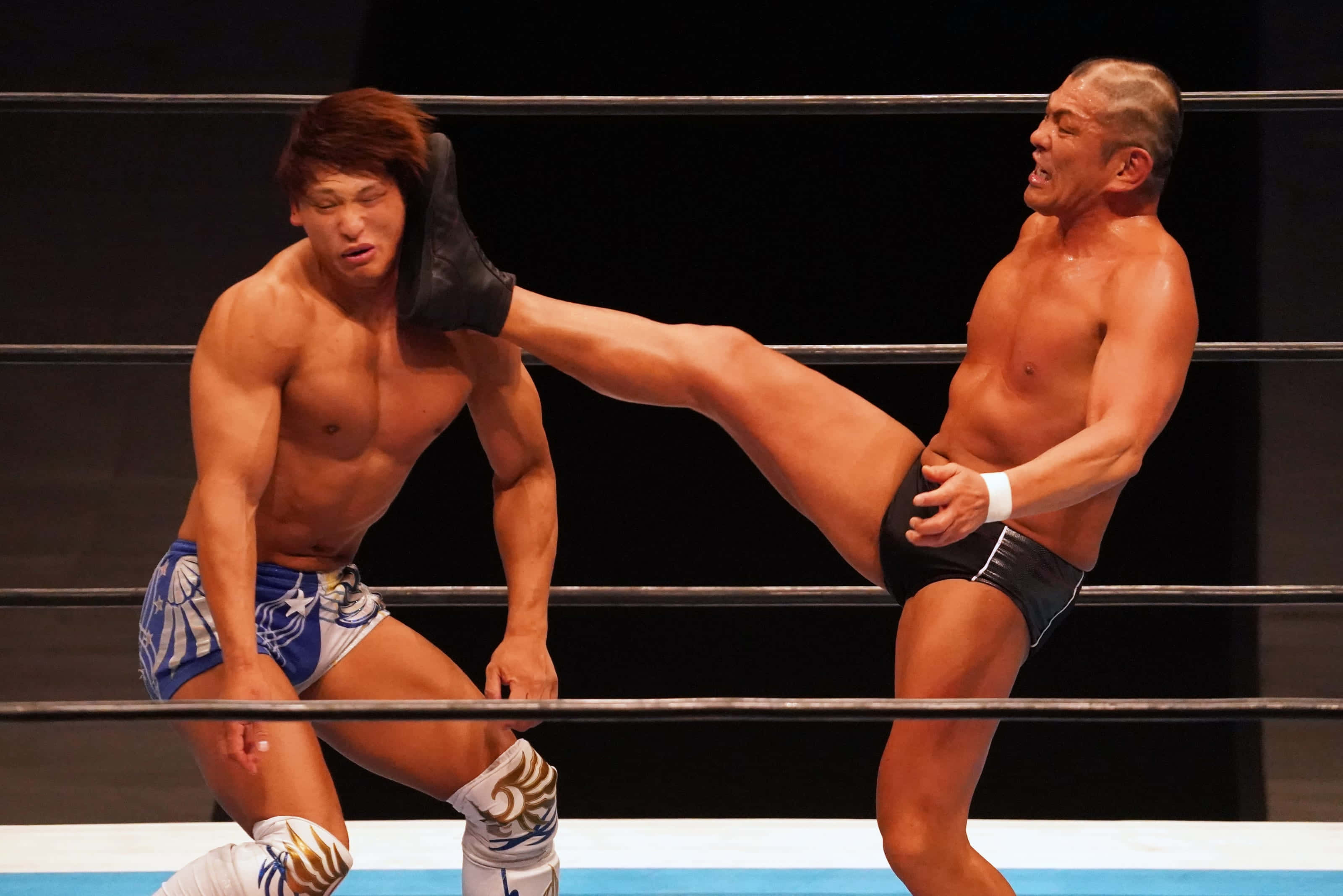 Minoru Suzuki 桌布 壁紙 — 兩名肌肉發達的摔角手在擂台上交鋒，其中一人在明亮的場館燈光下向對方的頭部踢出一記高踢。