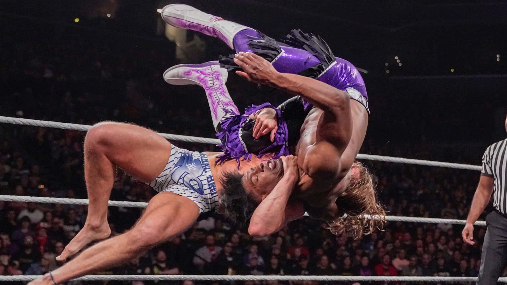 WWE 桌布 壁紙 — 兩名肌肉發達的摔角手在擂台上半空中糾纏，裁判在場館燈光下從旁觀看。