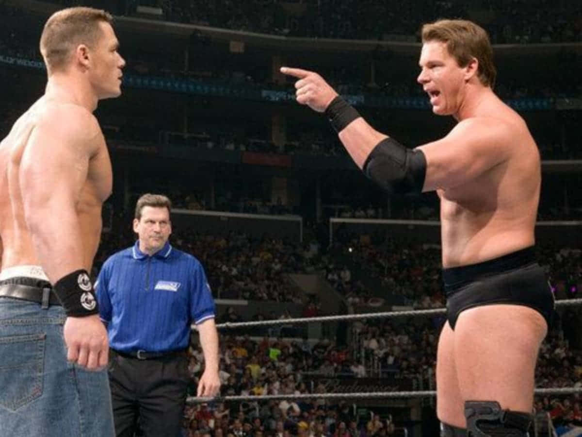 WWE 桌布 壁紙 — 兩名肌肉發達的摔角手在擂台上對峙，中間有裁判，觀眾緊張地觀看。