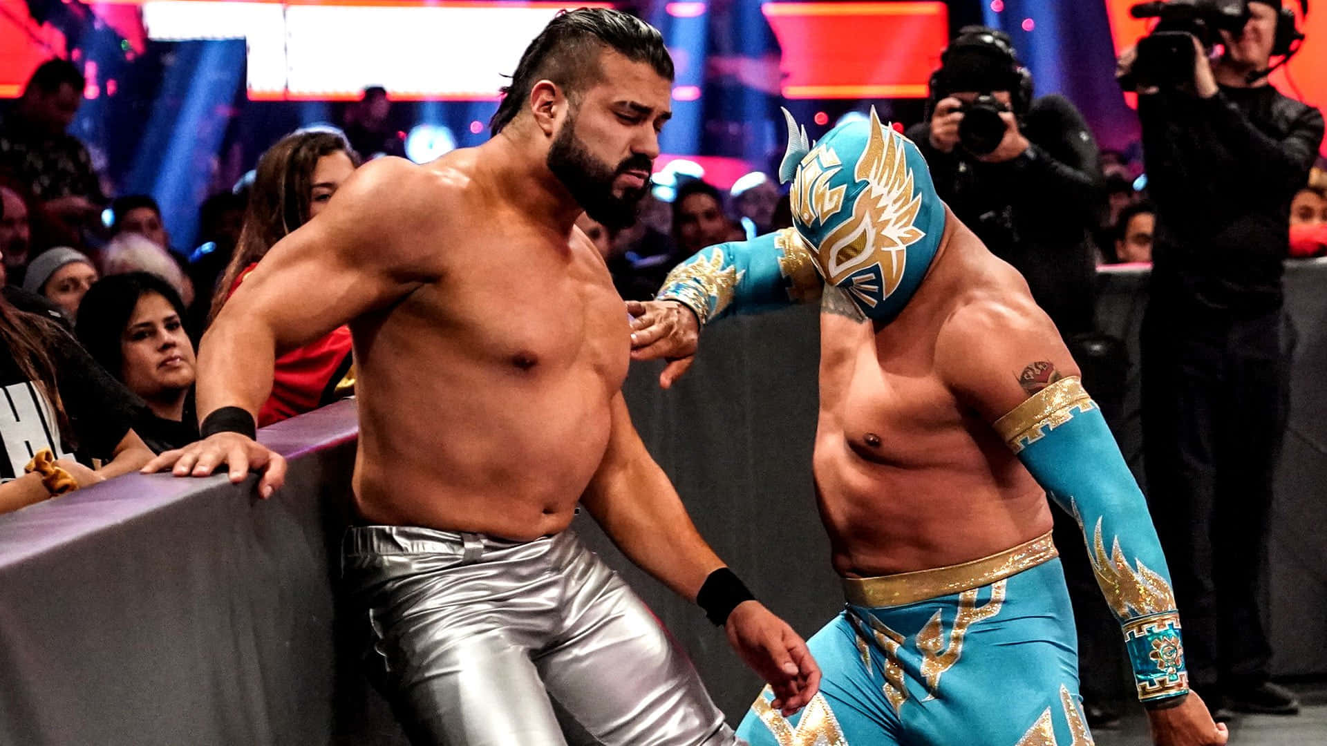 WWE 桌布 壁紙 — 兩名肌肉發達的摔角手在擂台上對峙，一人赤裸上身穿著銀色褲子，另一人穿著藍金色自由式摔角裝備。