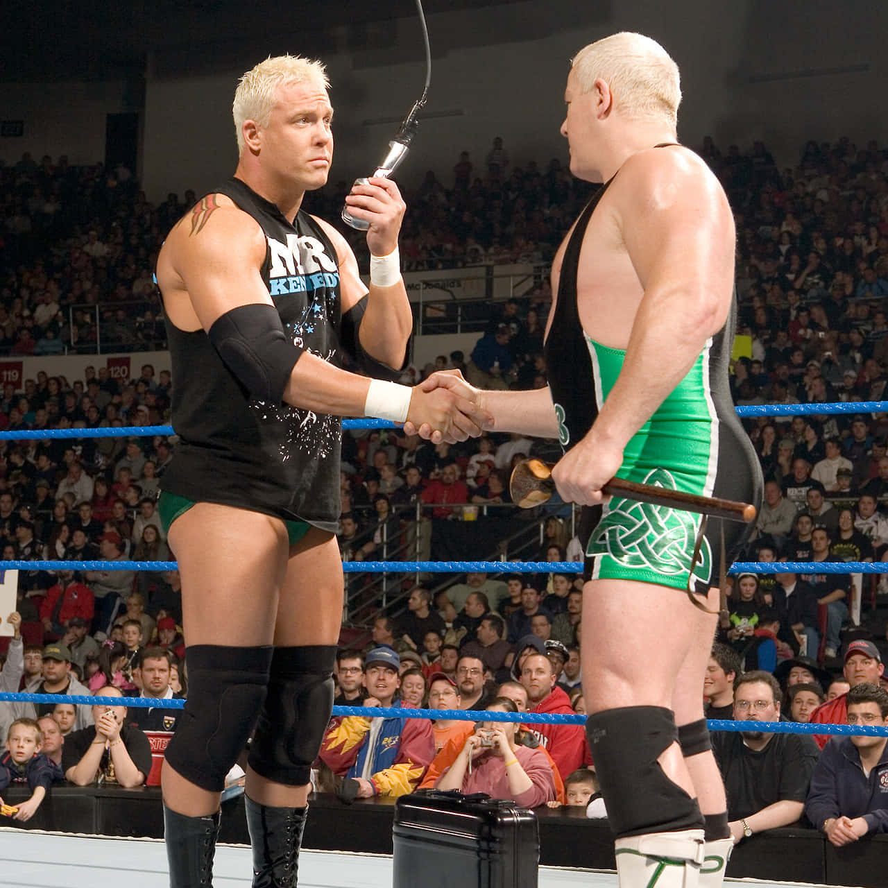 WWE 桌布 壁紙 — 兩名肌肉發達的摔角手在擂台上握手，一人拿著麥克風，另一人拿著球棒，觀眾在觀看。