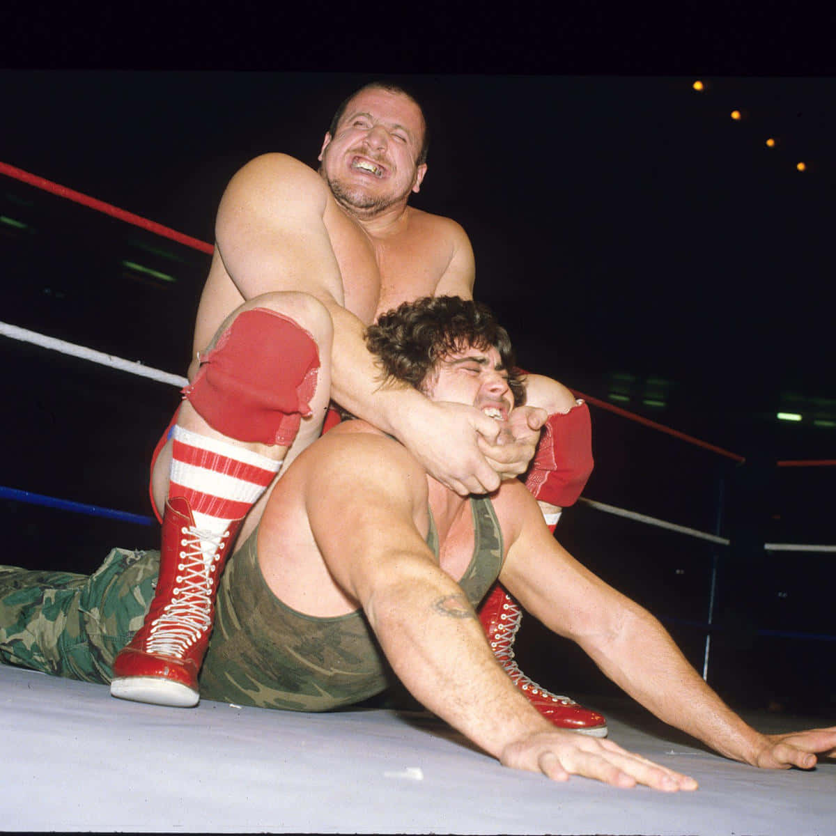 WWE 桌布 壁紙 — 兩名肌肉發達的摔角手在擂台上激烈纏鬥，一人穿著紅色靴子和迷彩褲，另一人穿著綠色短褲。