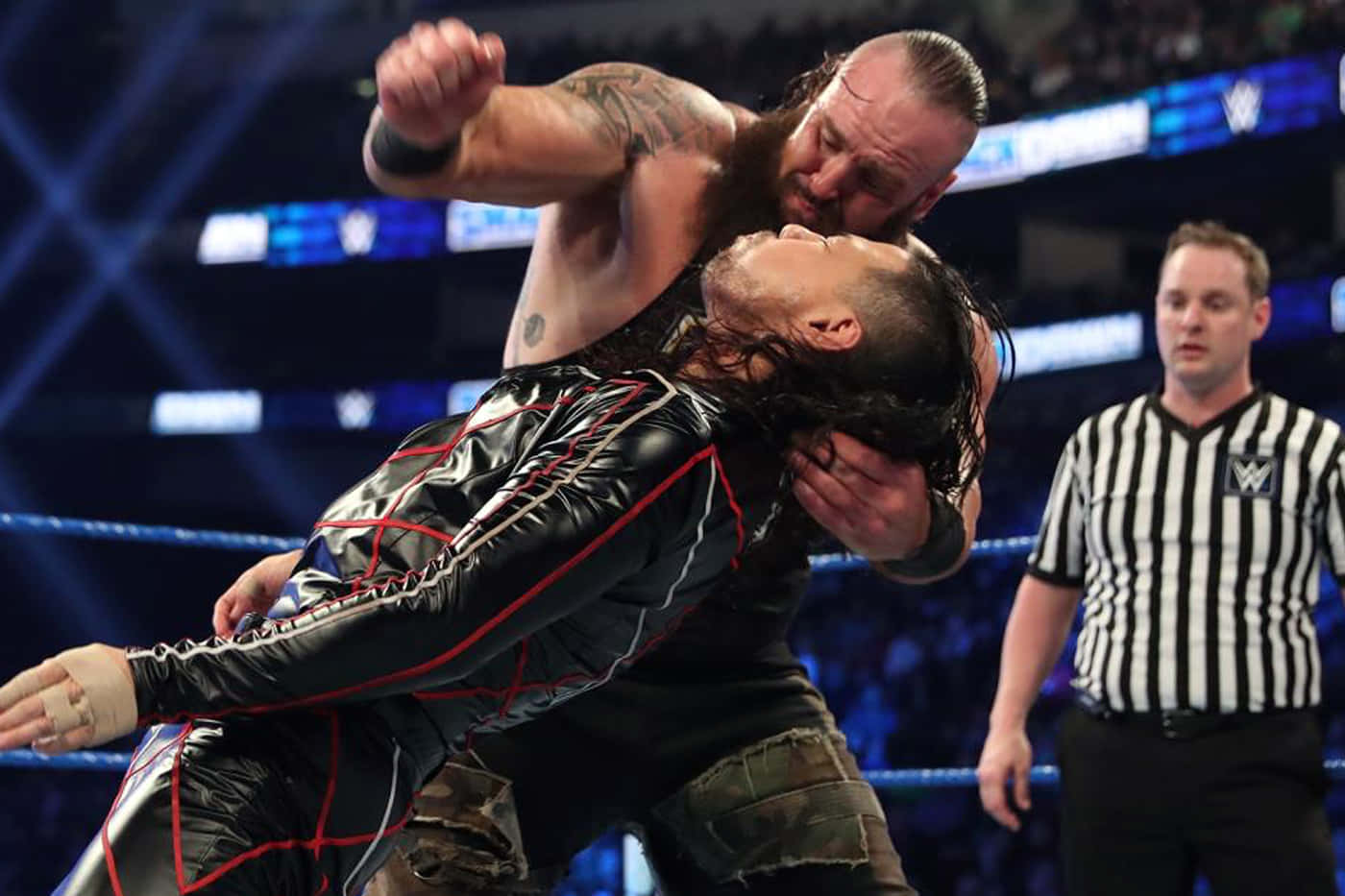 WWE 桌布 壁紙 — 兩名肌肉發達的摔角手在明亮的場館燈光下於擂台上激烈纏鬥，裁判在旁觀看。