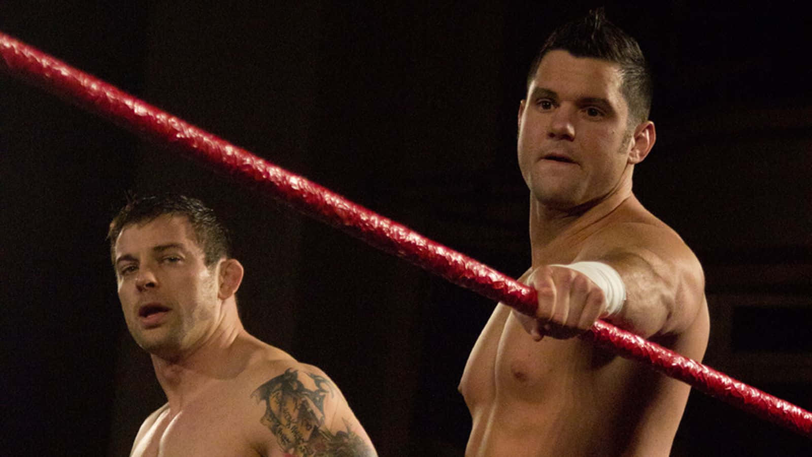 WWE 桌布 壁紙 — 兩名肌肉發達的摔角手在昏暗的場館中進行激烈比賽時抓住紅色擂台繩索。