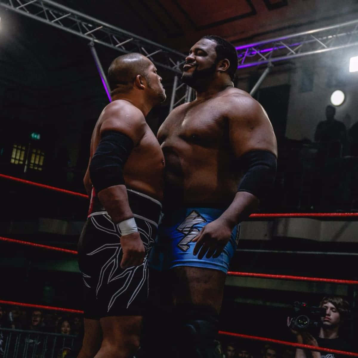 WWE 桌布 壁紙 — 兩名肌肉發達的摔角手在昏暗的擂台上，在戲劇性的紫色燈光下對峙，背景有觀眾。