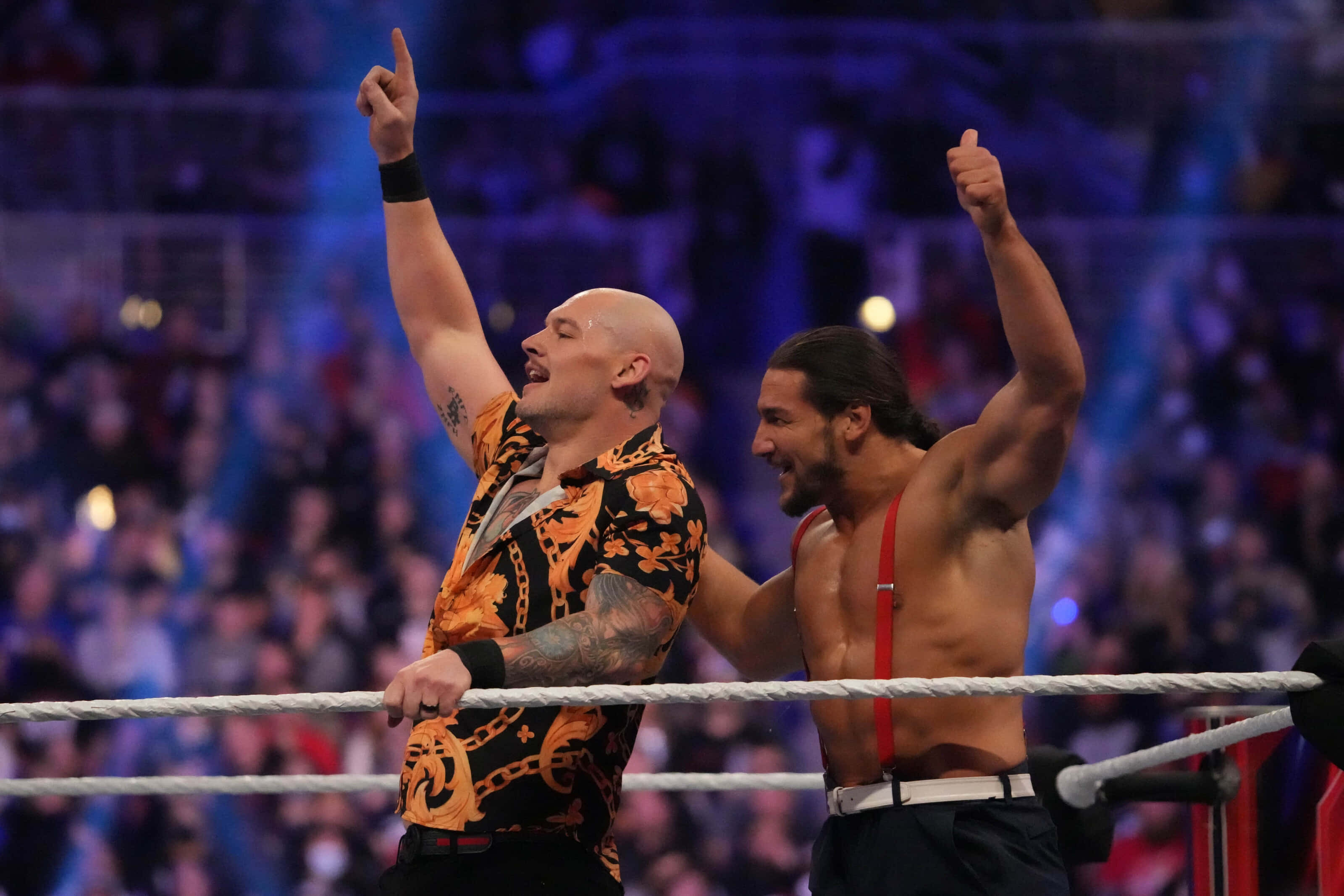 WWE 桌布 壁紙 — 兩名肌肉發達的摔角手在燈光明亮的擂台上慶祝勝利，背景有歡呼的粉絲。