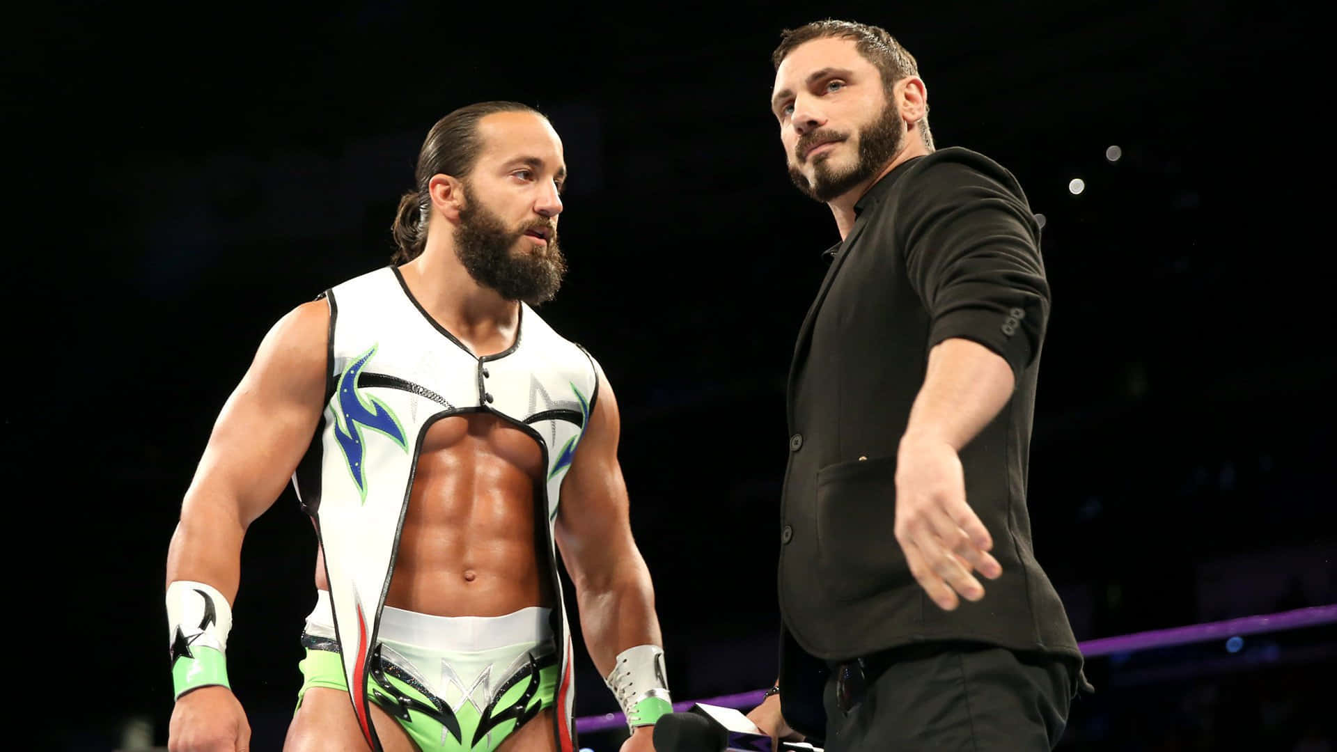 WWE 桌布 壁紙 — 兩名肌肉發達的摔角手在燈光明亮的擂台上對峙，一人身穿彩色裝備，另一人穿著黑色西裝。