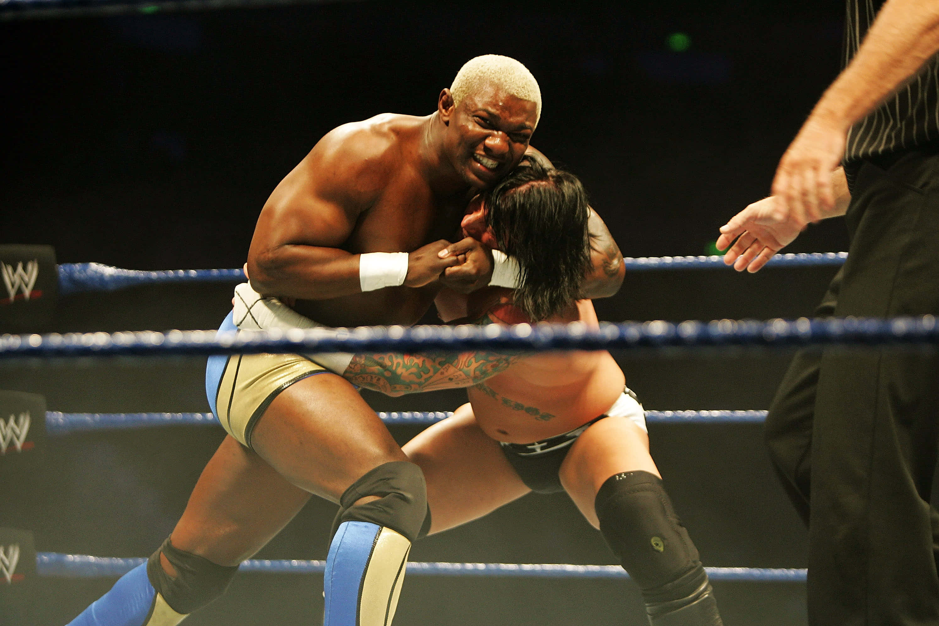 WWE 桌布 壁紙 — 兩名肌肉發達的摔角手在燈光明亮的擂台上激烈纏鬥，附近有一名裁判。