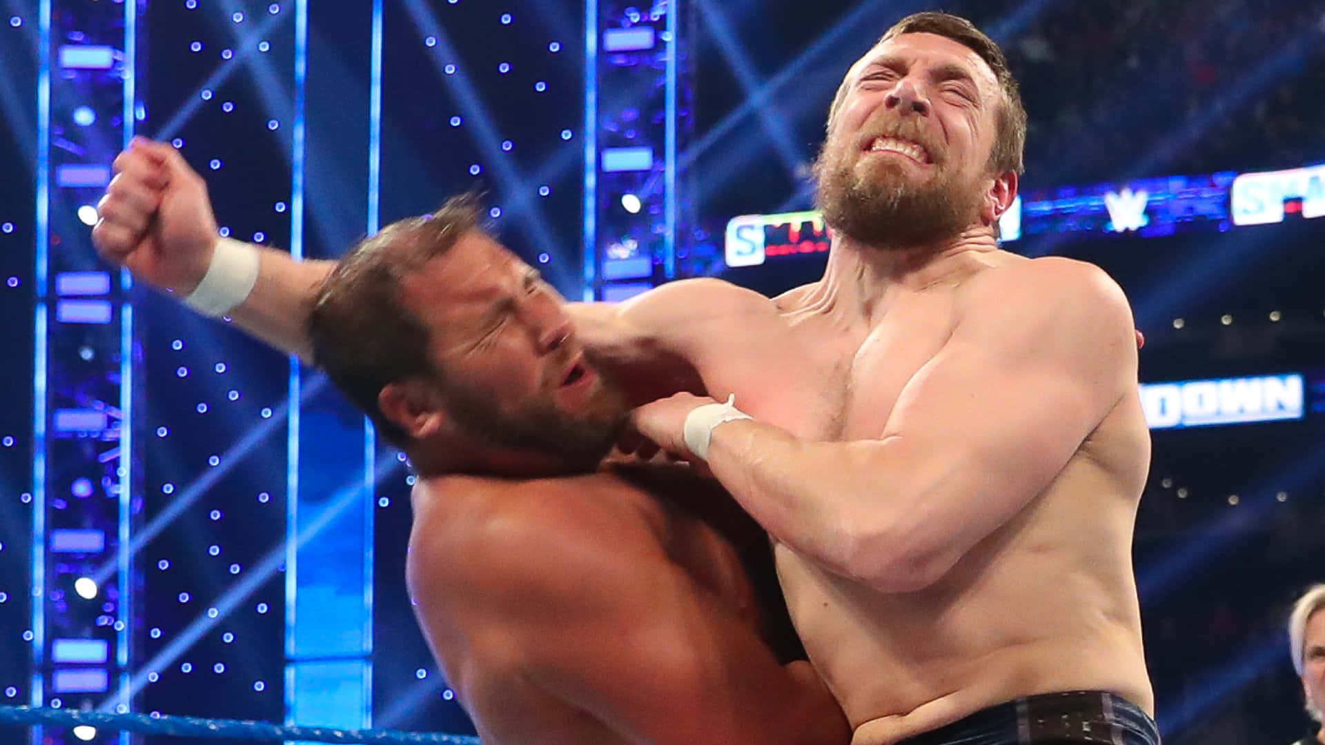 WWE 桌布 壁紙 — 兩名肌肉發達的摔角手在燈光明亮的擂台上激烈纏鬥，擂台上有藍色聚光燈和場館標誌。