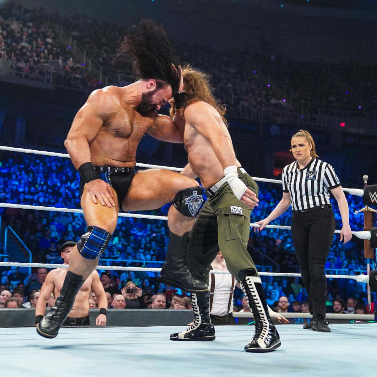WWE 桌布 壁紙 — 兩名肌肉發達的摔角手在燈光明亮的擂台上激烈糾纏，裁判在咆哮的人群旁密切注視。